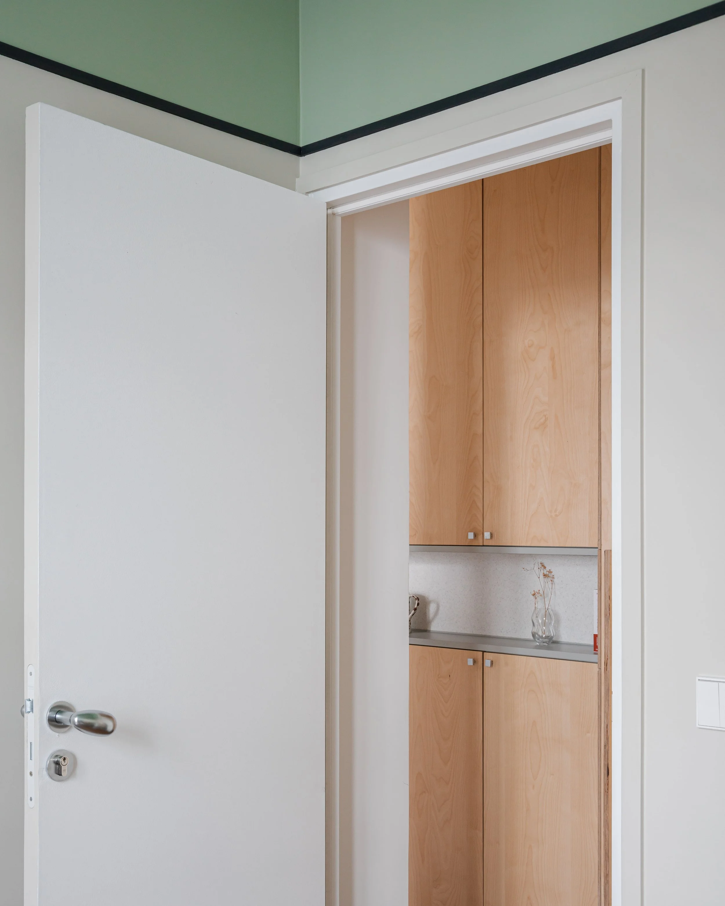 Une porte de chambre entrouverte donnant sur un placard en bois avec des portes en palette, contenant un vase avec des fleurs séchées et un petit objet rouge.