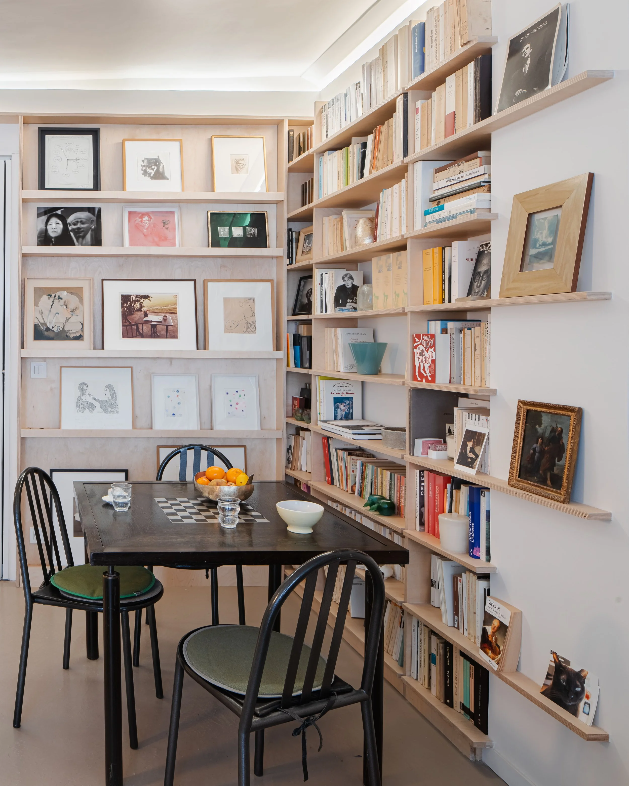 Une bibliothèque remplie de livres, avec des œuvres d'art encadrées et un tableau de jeu d'échecs, accompagnée d'une table avec des fruits et quelques objets décoratifs.