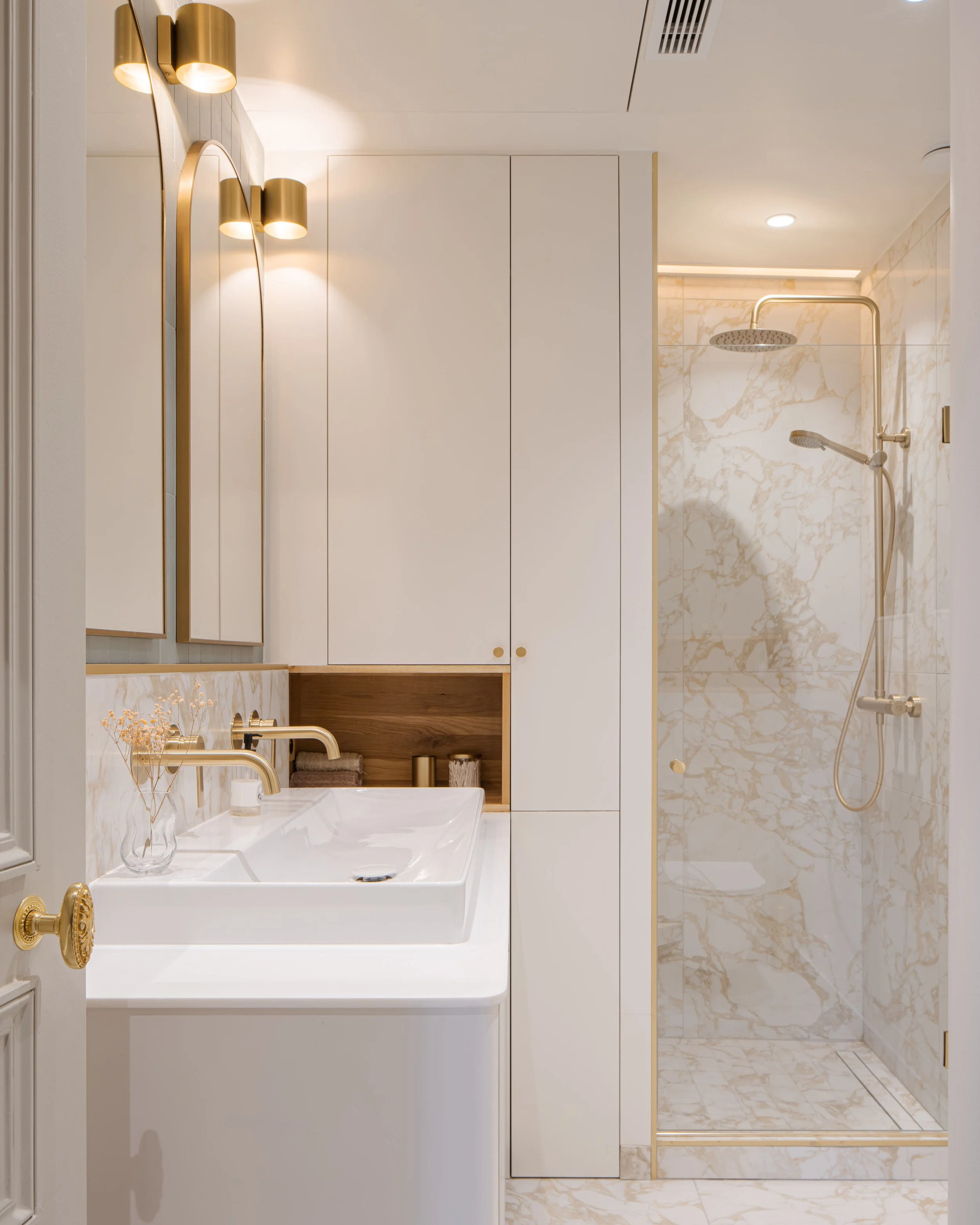 Salles de bain élégante avec lavabo blanc, grand miroir, armoires blanches, éclairage en or mat, espace douche avec murs en marbre beige, douchette haute et flexible.