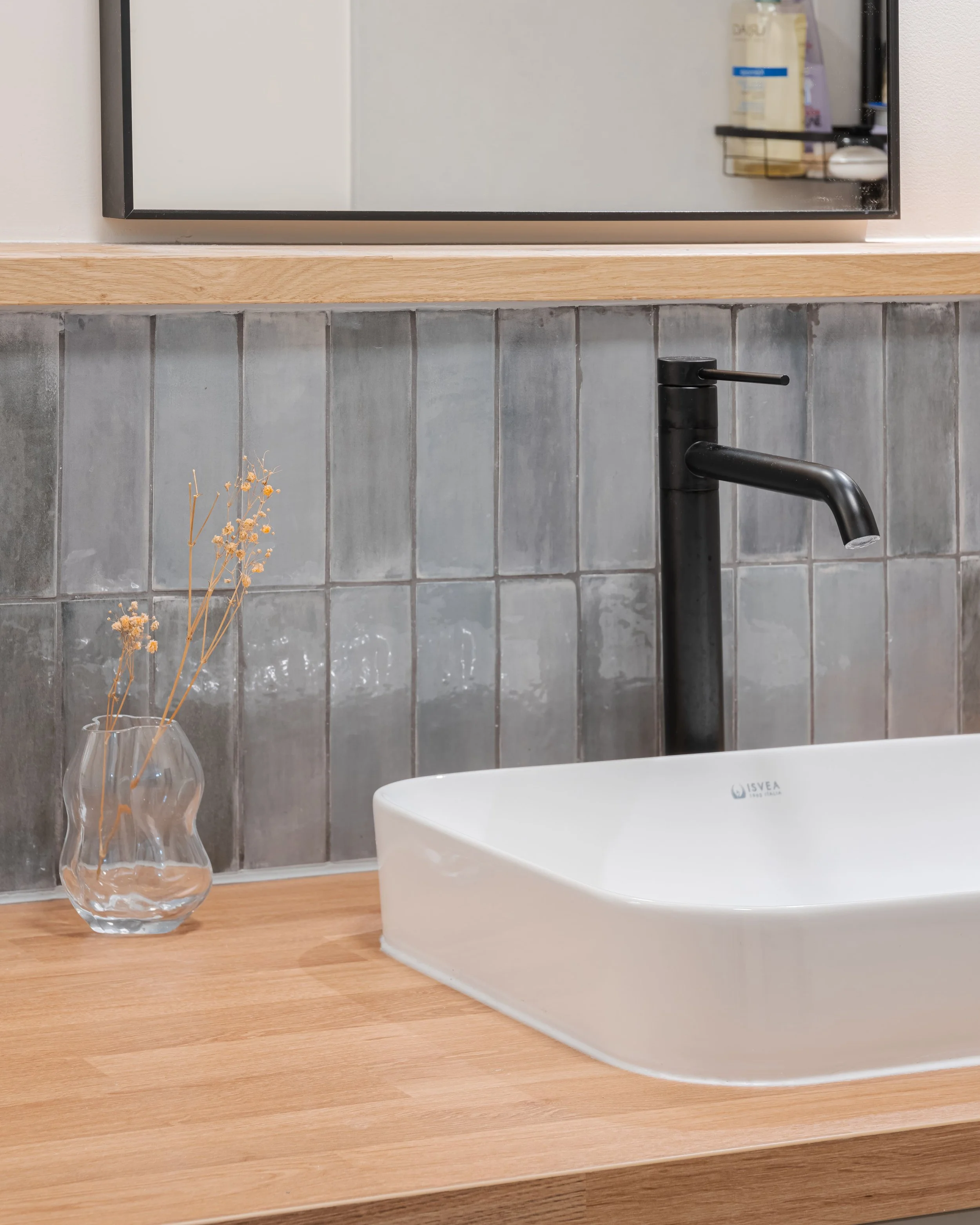Lavabo blanc avec robinet noir, vase en verre avec des herbes séchées, miroir au-dessus, carrelage gris derrière, plan de bois autour du lavabo dans une salle de bain.
