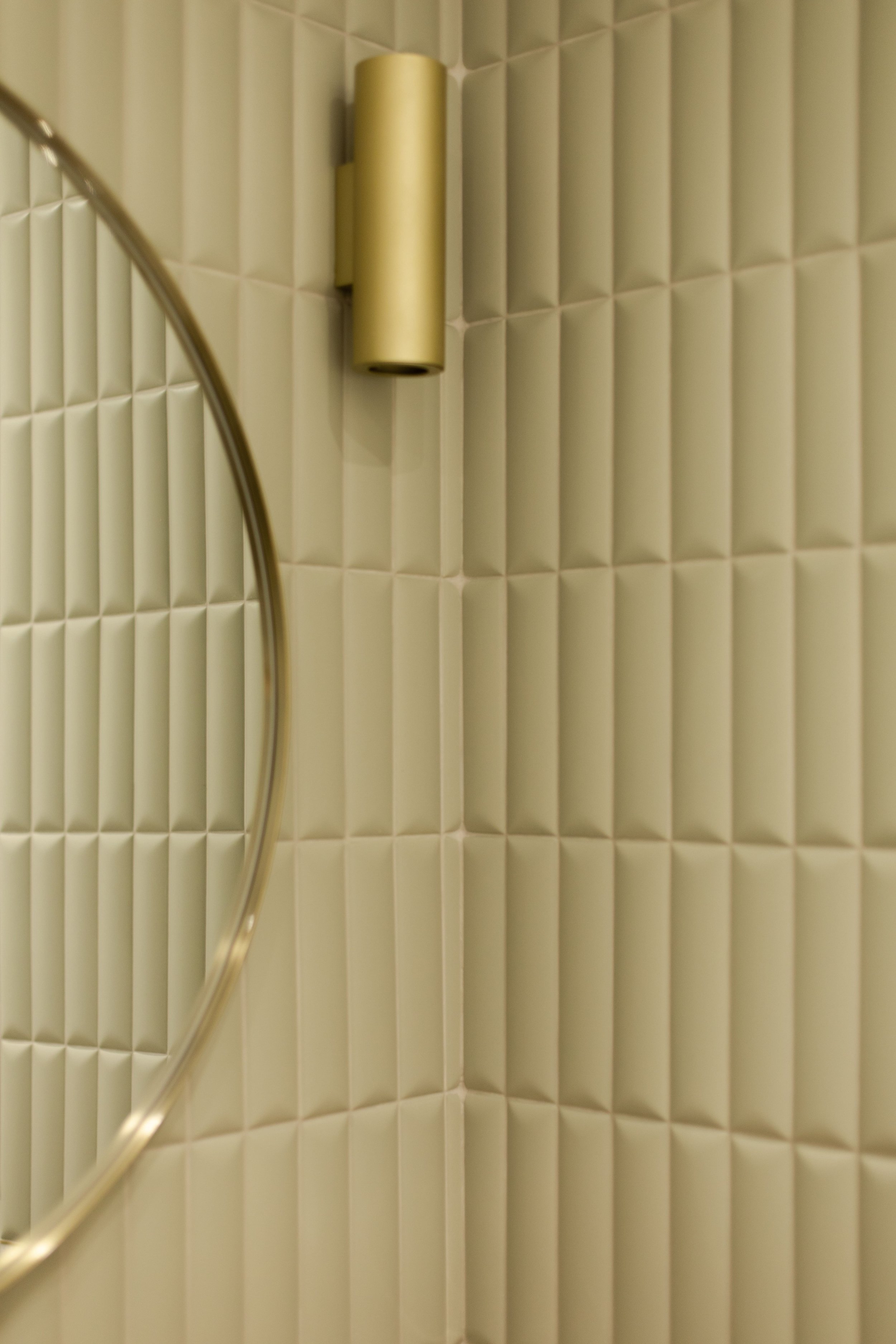 Mur en carreaux 3D beige avec un miroir rond partiellement visible et une applique murale jaune.