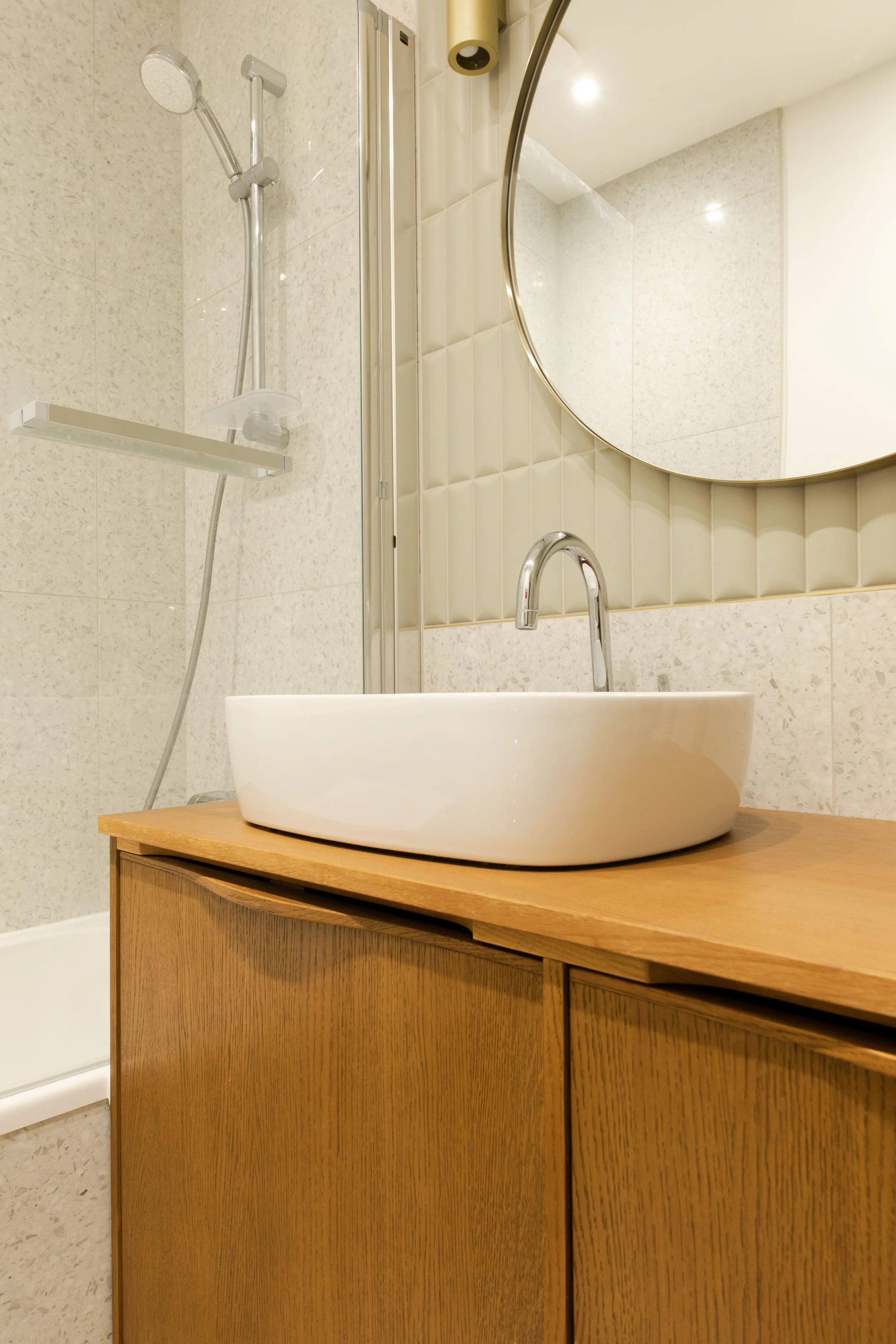 Salle de bain avec un lavabo rond blanc sur une armoire en bois, un miroir rond au-dessus, une douche avec une tête de douche et un siège en arrière-plan.
