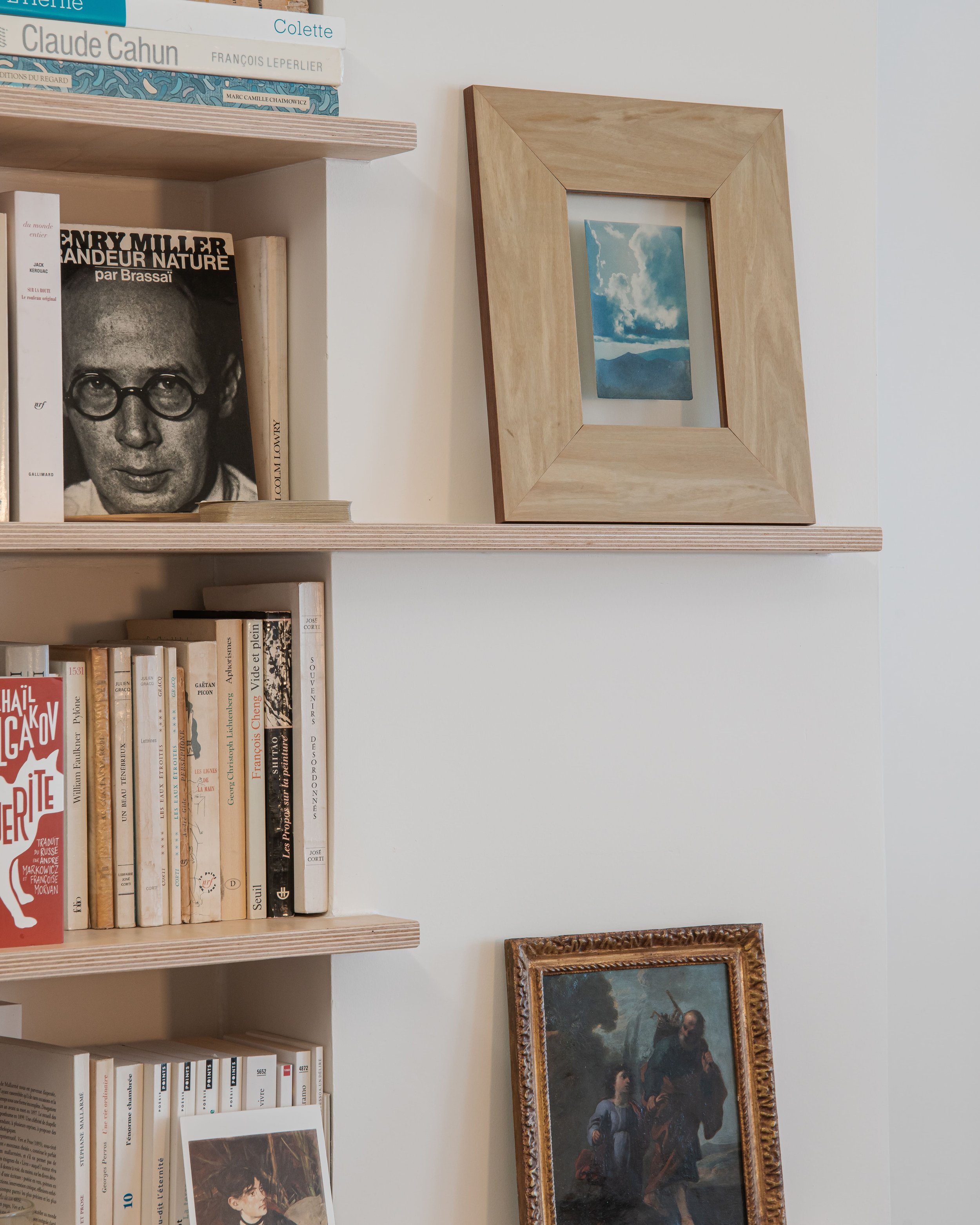 Une étagère en bois avec des livres, une affiche en noir et blanc d'Henry Miller, une petite peinture de ciel avec clouds, et un tableau classique illustrant une scène d'un homme et une femme.