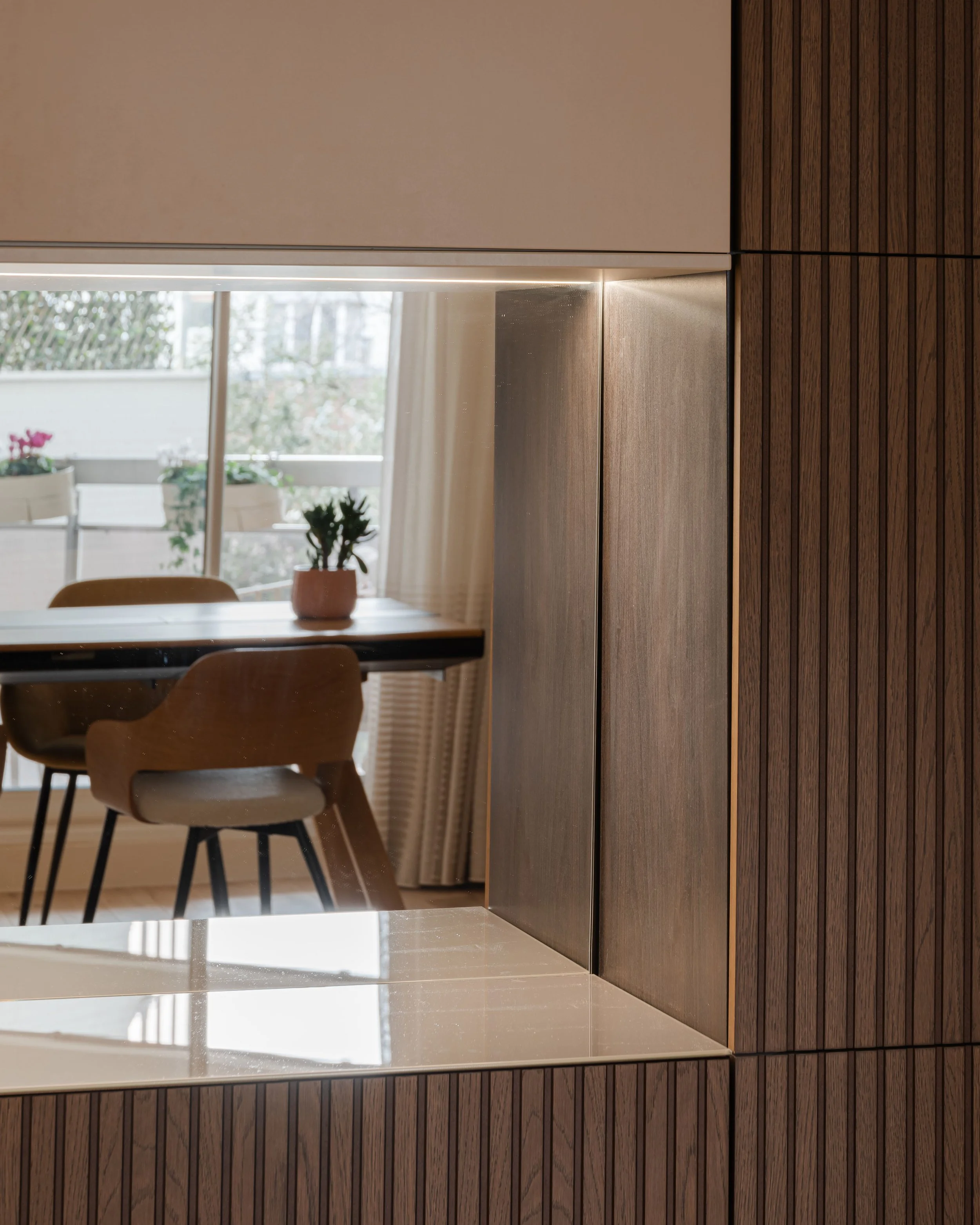 Vue partielle d'une pièce avec une table, des chaises en bois, et une fenêtre laissant entrer de la lumière naturelle, décoration intérieure moderne avec bois clair et accents en noir.