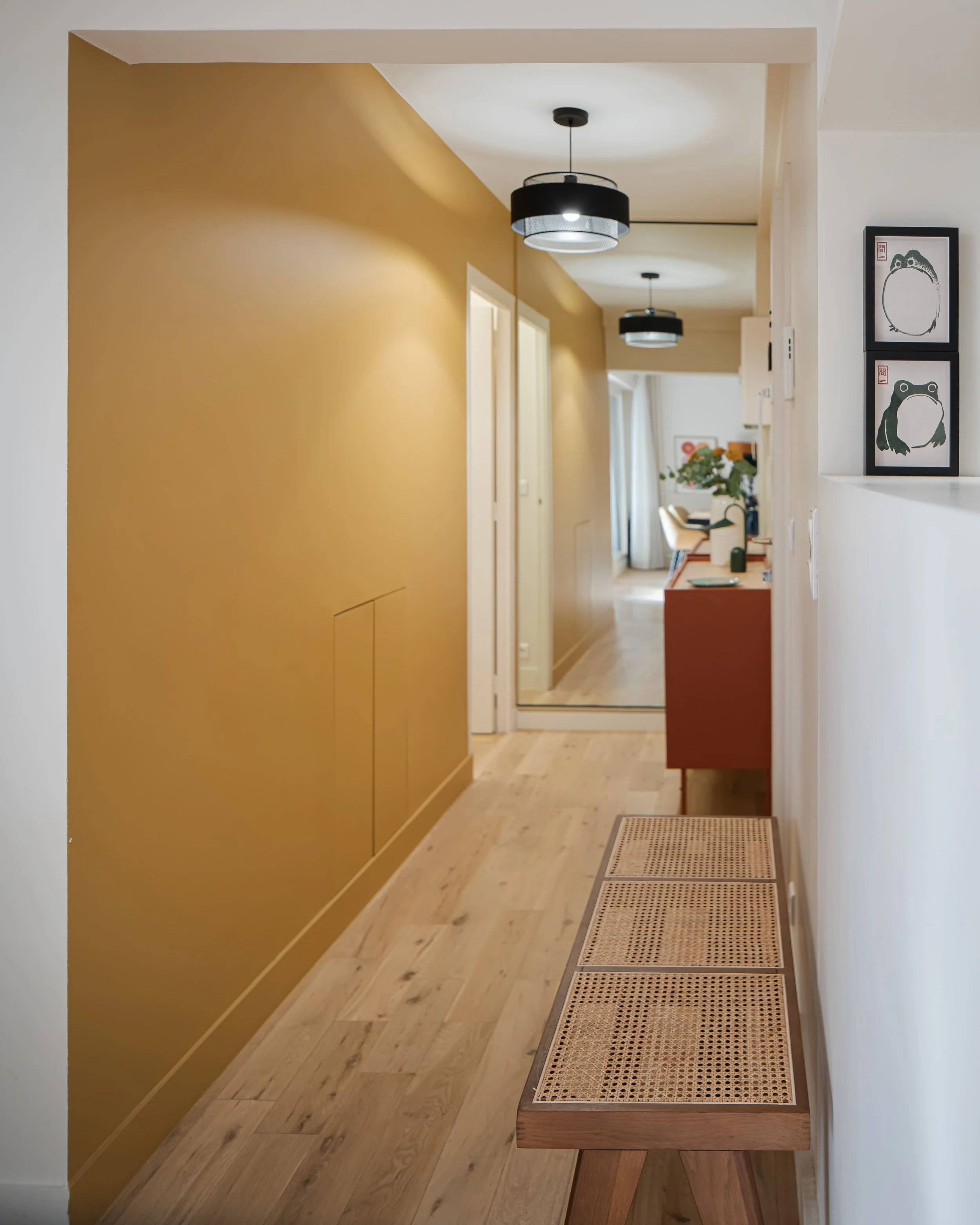 Couloir d'une maison avec mur jaune à gauche, revêtement de sol en bois clair, lampes suspendues noires, dessin de grenouilles encadré à droite, mobilier décoratif à l'arrière.