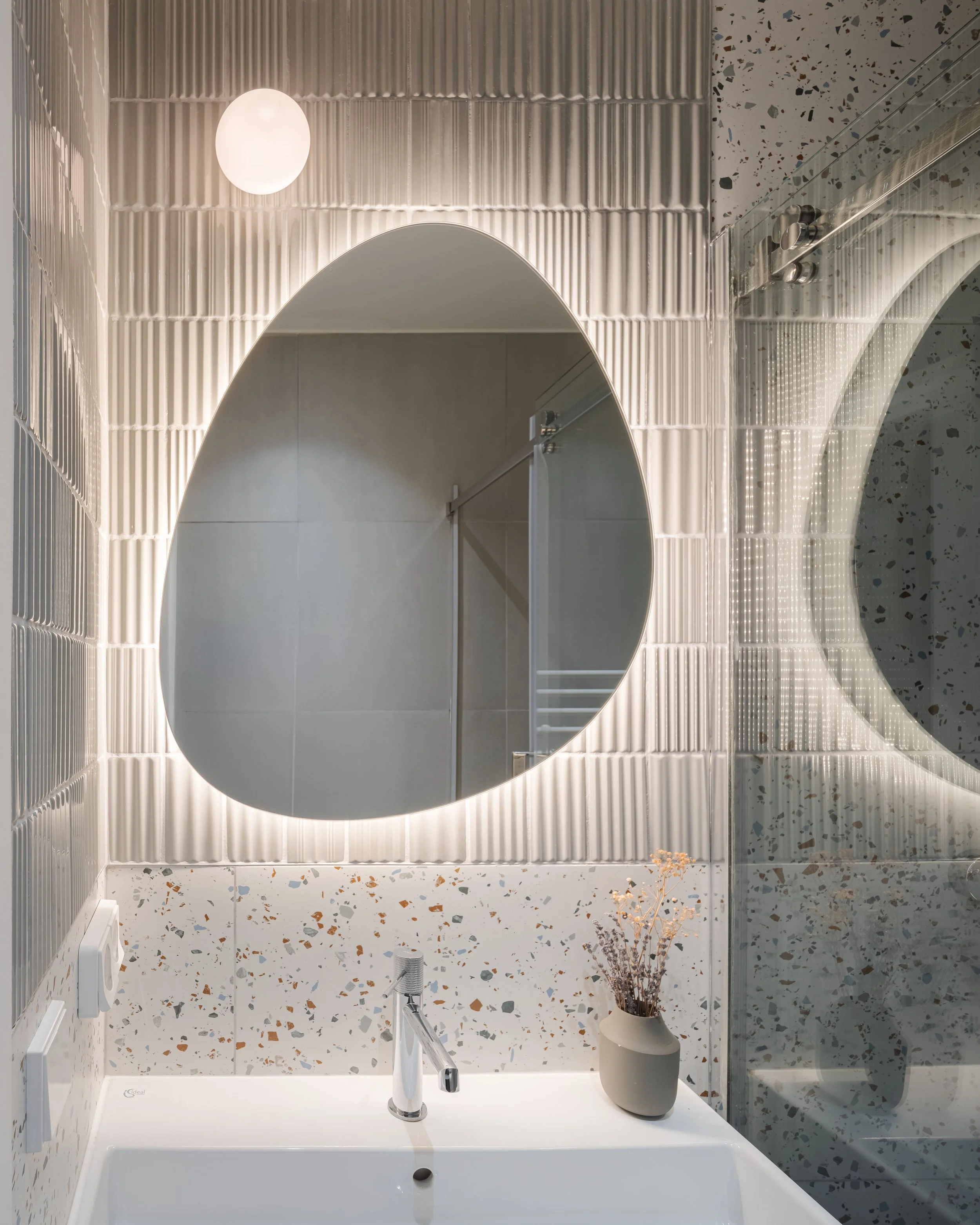 Miroir rond avec éclairage LED derrière, lavabo blanc avec robinet, vase beige avec fleurs séchées, carrelage murale gris et terrazzo au sol, lumière ronde au plafond.