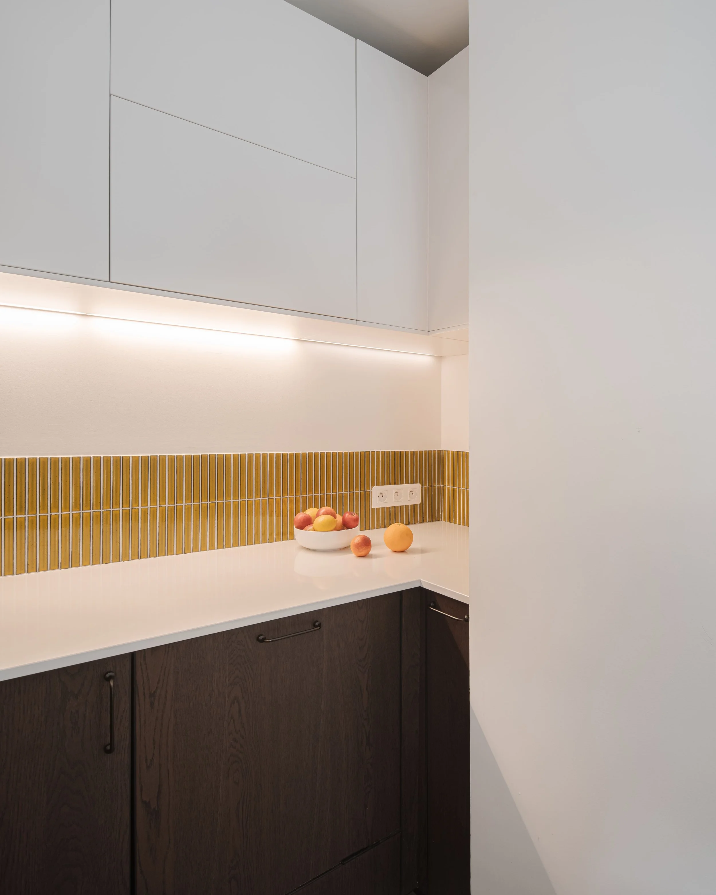 Coin cuisine avec des armoires blanches, un plan de travail blanc, une bande de carreaux de métro jaunes, une bowl de fruits, et une prise électrique.