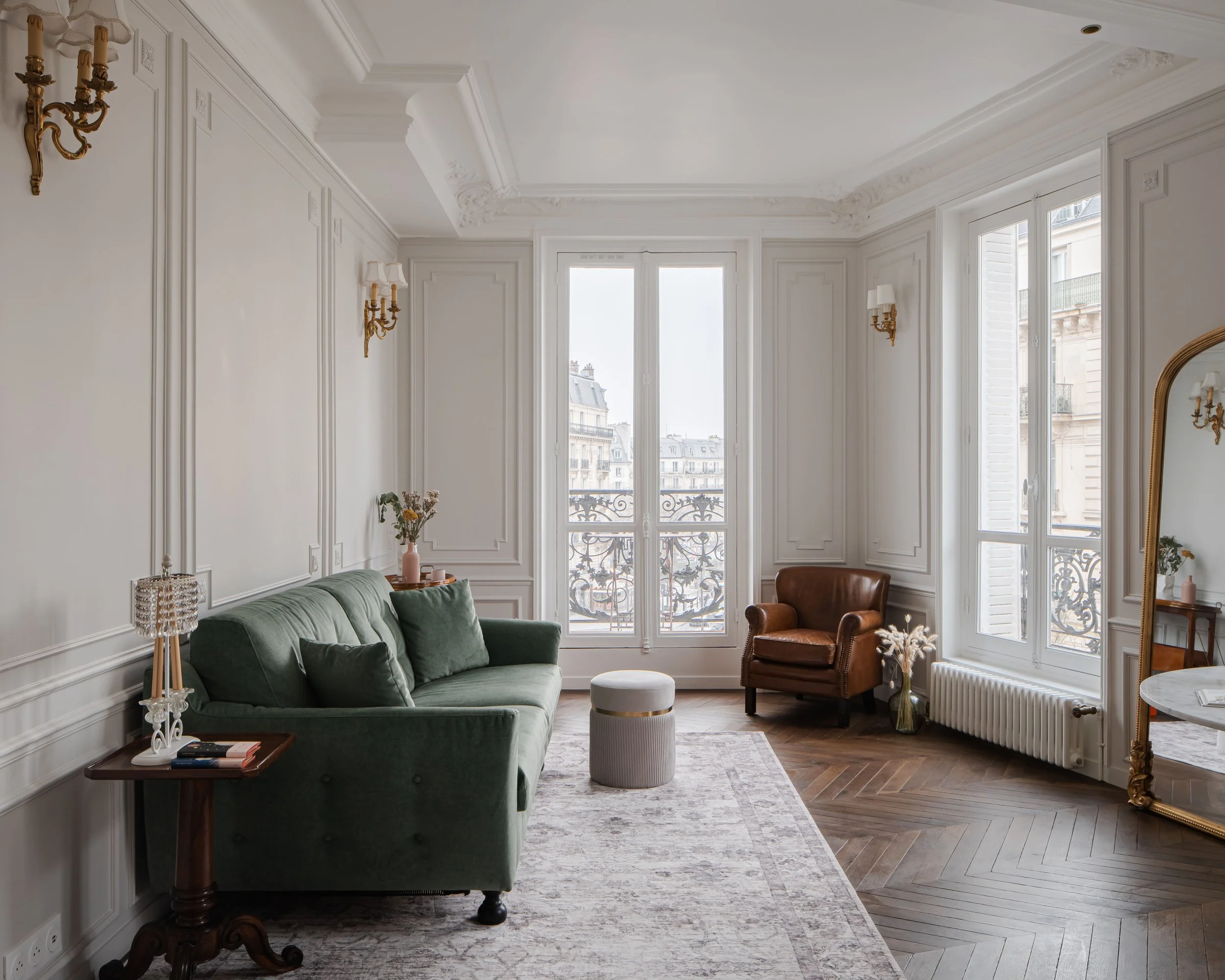 Salon lumineux avec canapé vert, fauteuil en cuir brun, grand miroir doré, fenêtres avec vue sur bâtiment parisien, décor classique avec moulures et lampes en or.