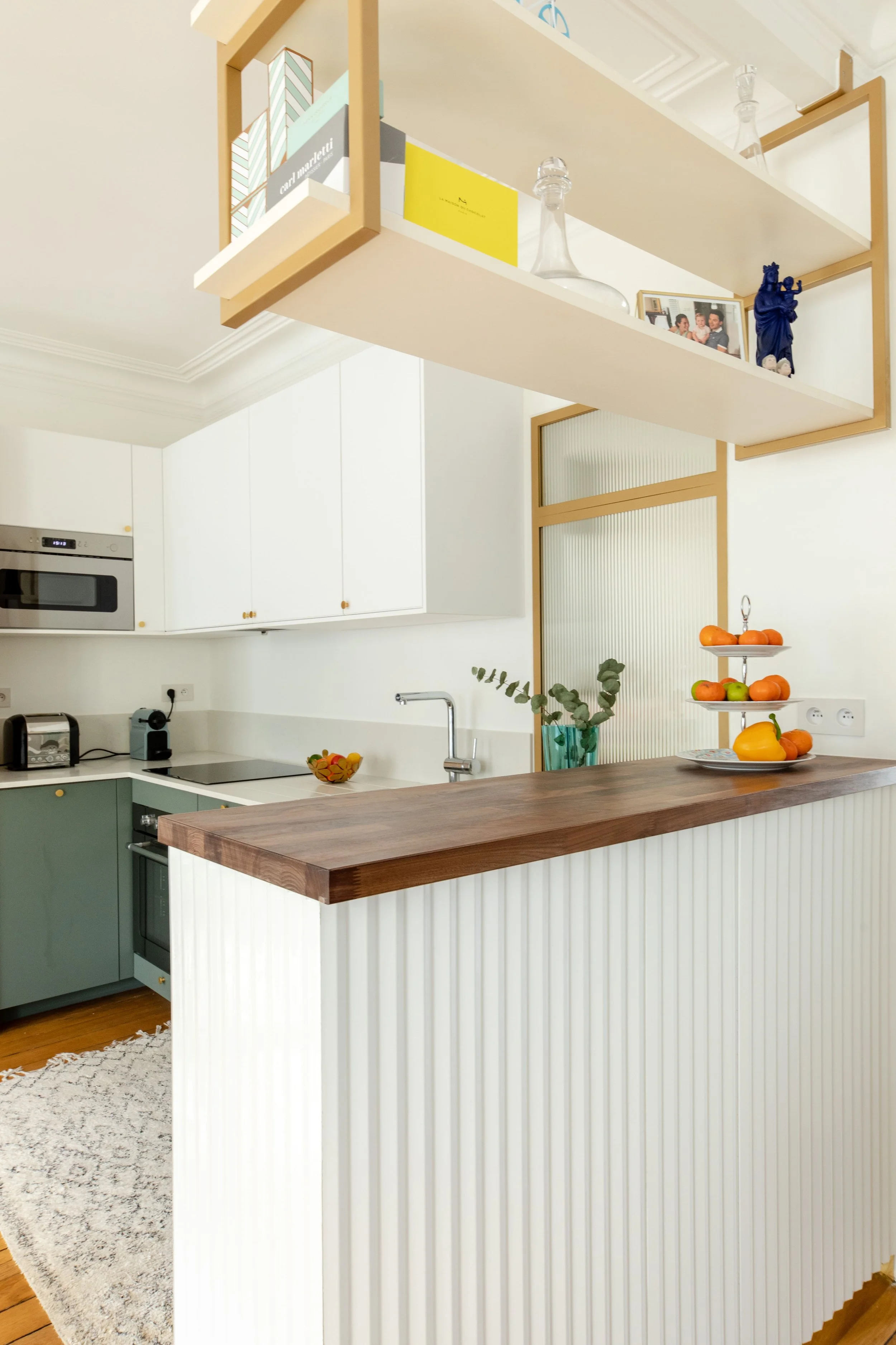 Cuisine moderne avec comptoir en bois, étagère suspendue avec livres, décorations et photo, murs blancs, armoire en deux couleurs, plantes et fruits en décoration.