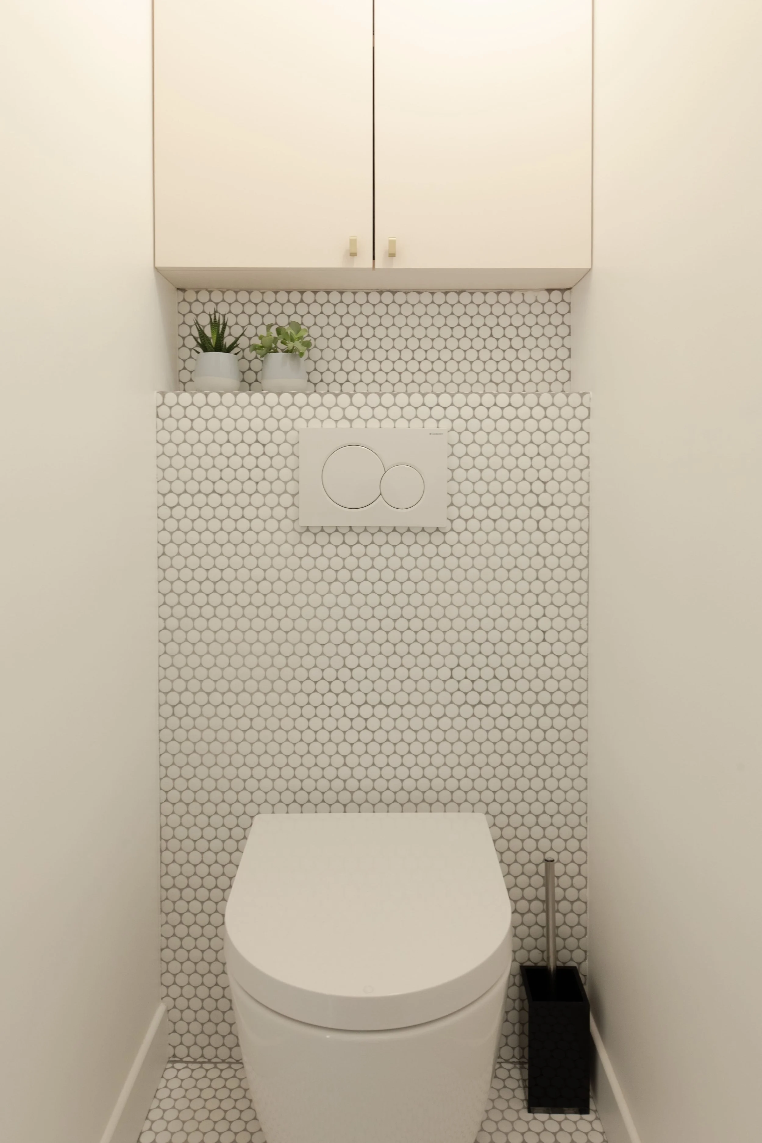 Toilettes modernes avec un mur en mosaïque hexagonale blanche, deux pots de fleurs en haut à gauche, un flusher encastré avec panneau de commande circulaire, un chapelet noir et une brosse à toilettes à droite.