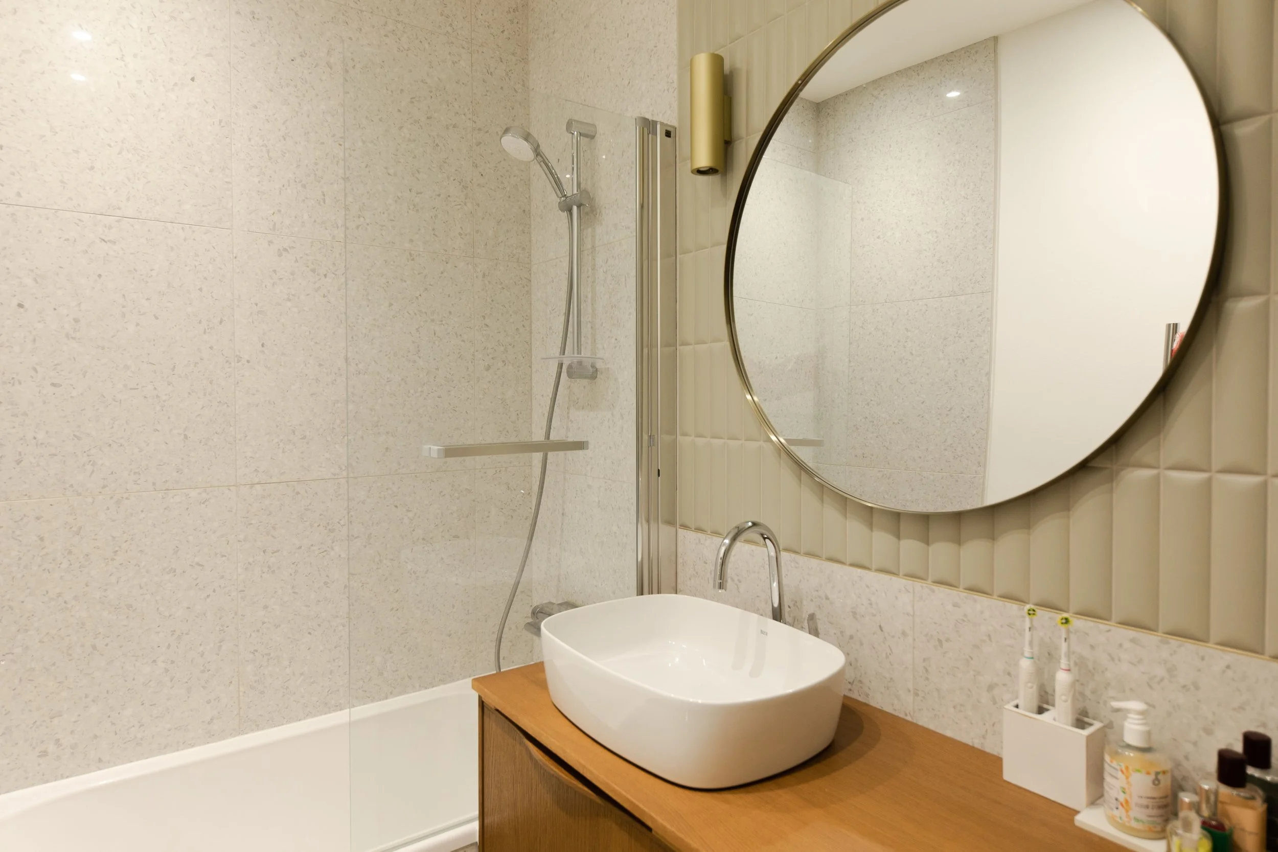 Salle de bain moderne avec un miroir rond, un lavabo blanc sur un meuble en bois, des brosses à dents, un savon et divers produits de toilette sur le comptoir, et une douche avec un pommeau et une paroi en verre.