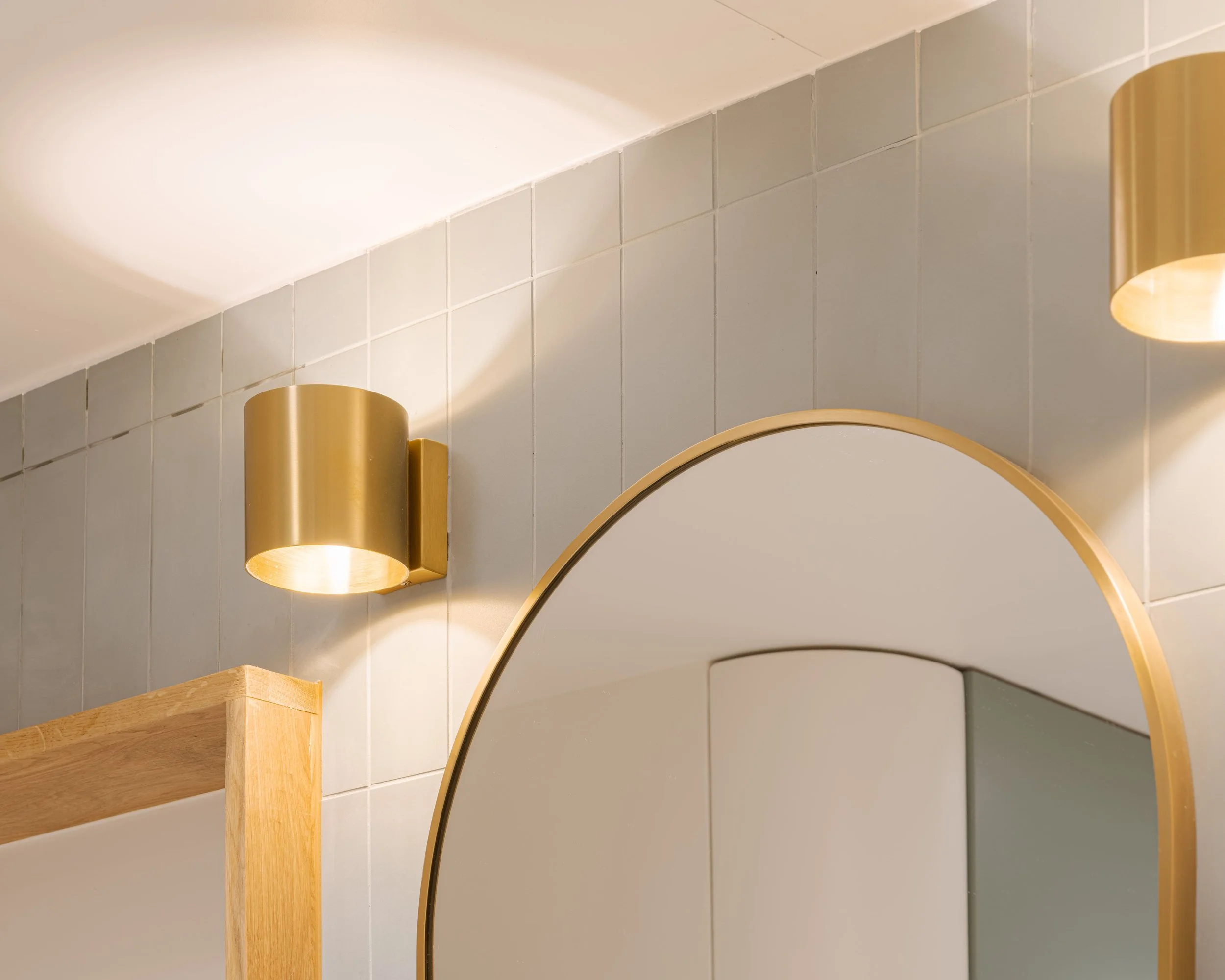Miroir rond avec cadre doré, lampe murale dorée, et mur en carreaux gris dans une salle de bain moderne.