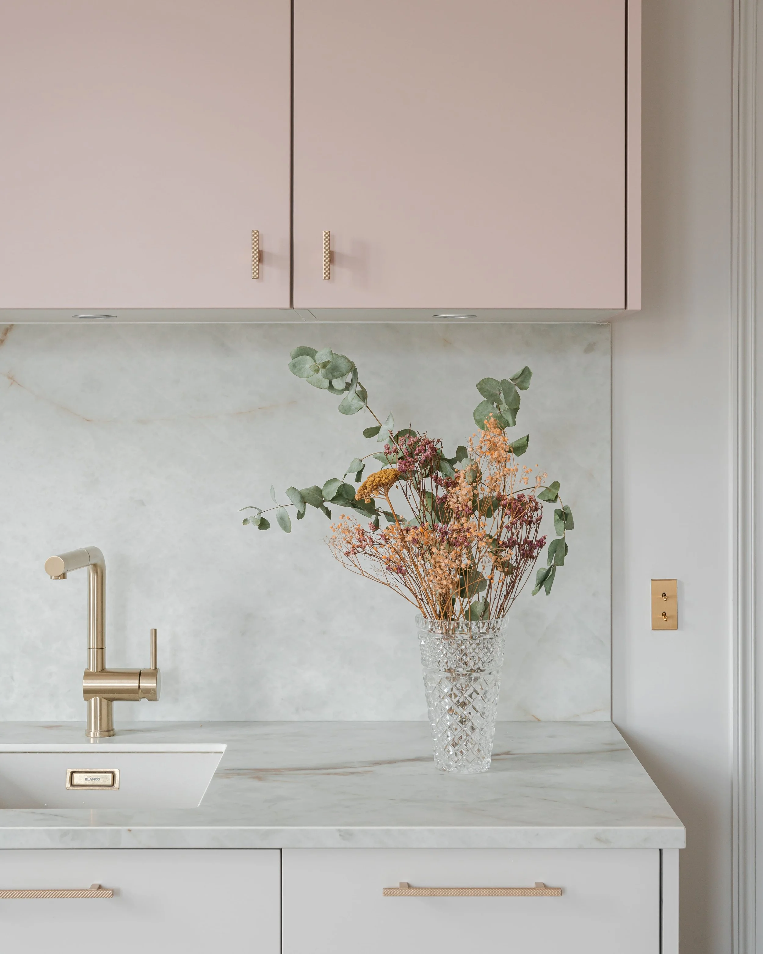 Une vasque en verre avec des fleurs séchées, un robinet doré, des armoires roses dans une cuisine moderne.