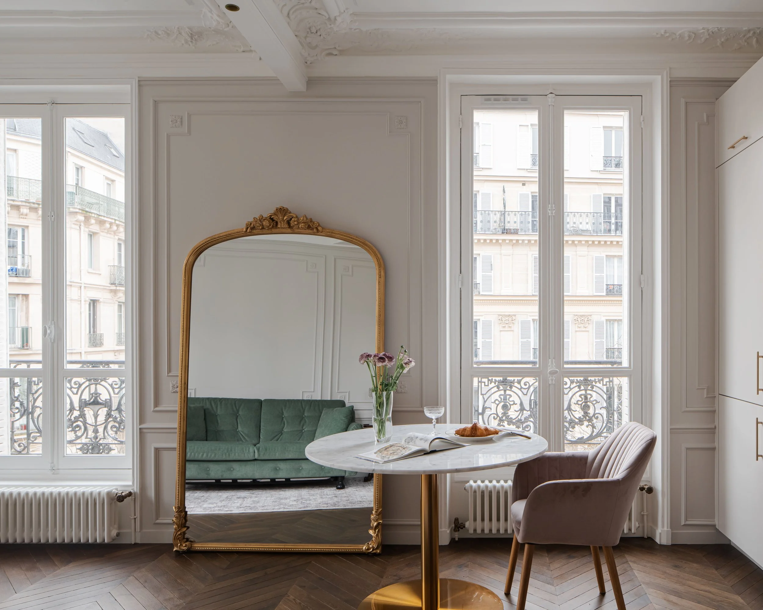 Salon intérieur avec grands fenêtres, miroir ancien, table ronde en marbre, fauteuil en velours rose, canapé vert, décor classique et parquet en bois.