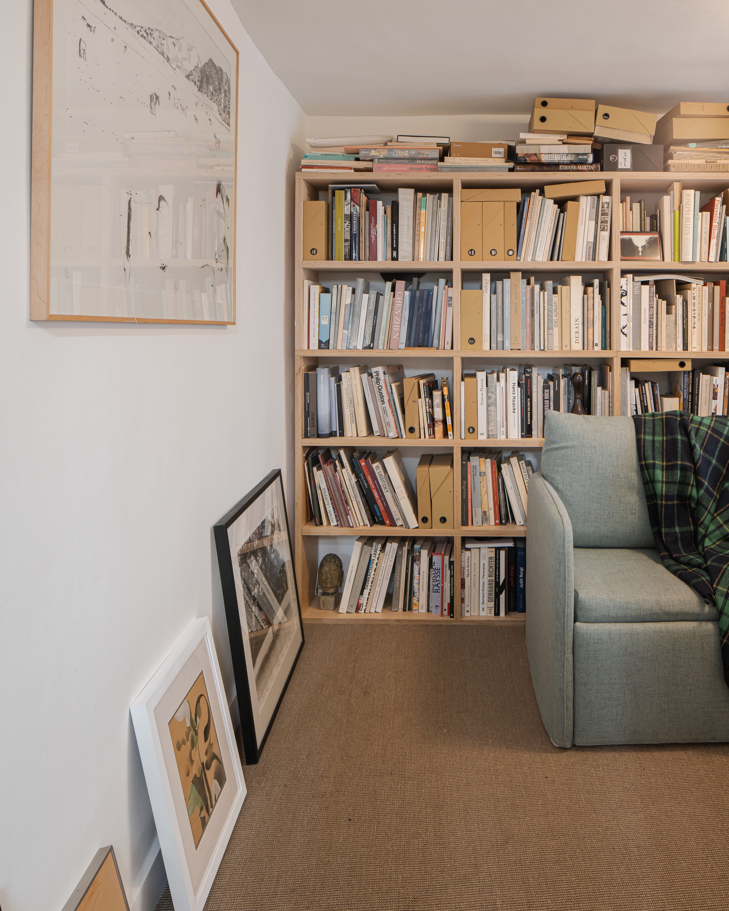 Une étagère en bois pleine de livres, avec des tableaux posés au sol, un canapé gris avec une couverture à carreaux, dans un salon ou une bibliothèque.