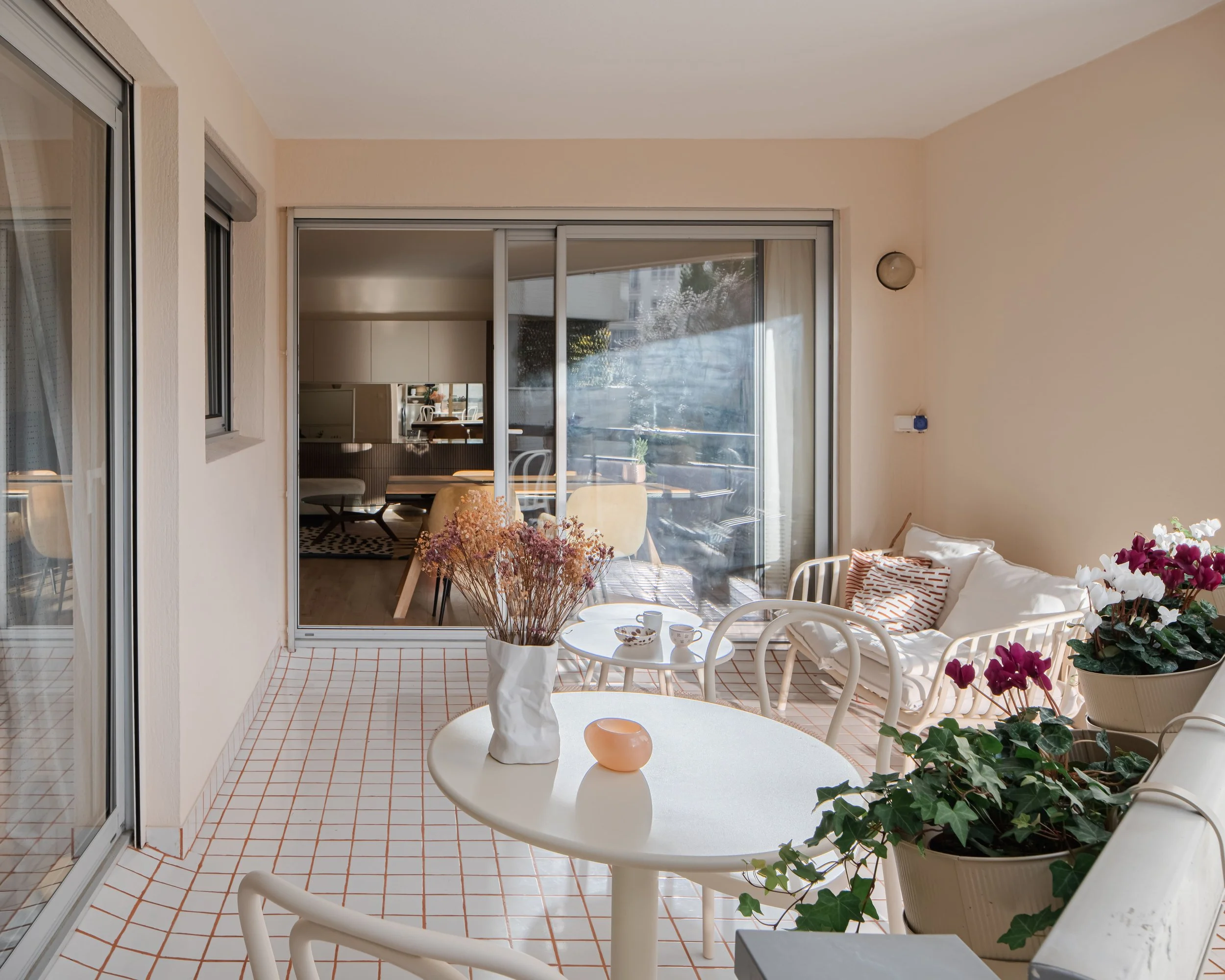 Balcon avec table, chaises, plantes en pot et coussins, vue sur intérieur salon avec salle à manger.