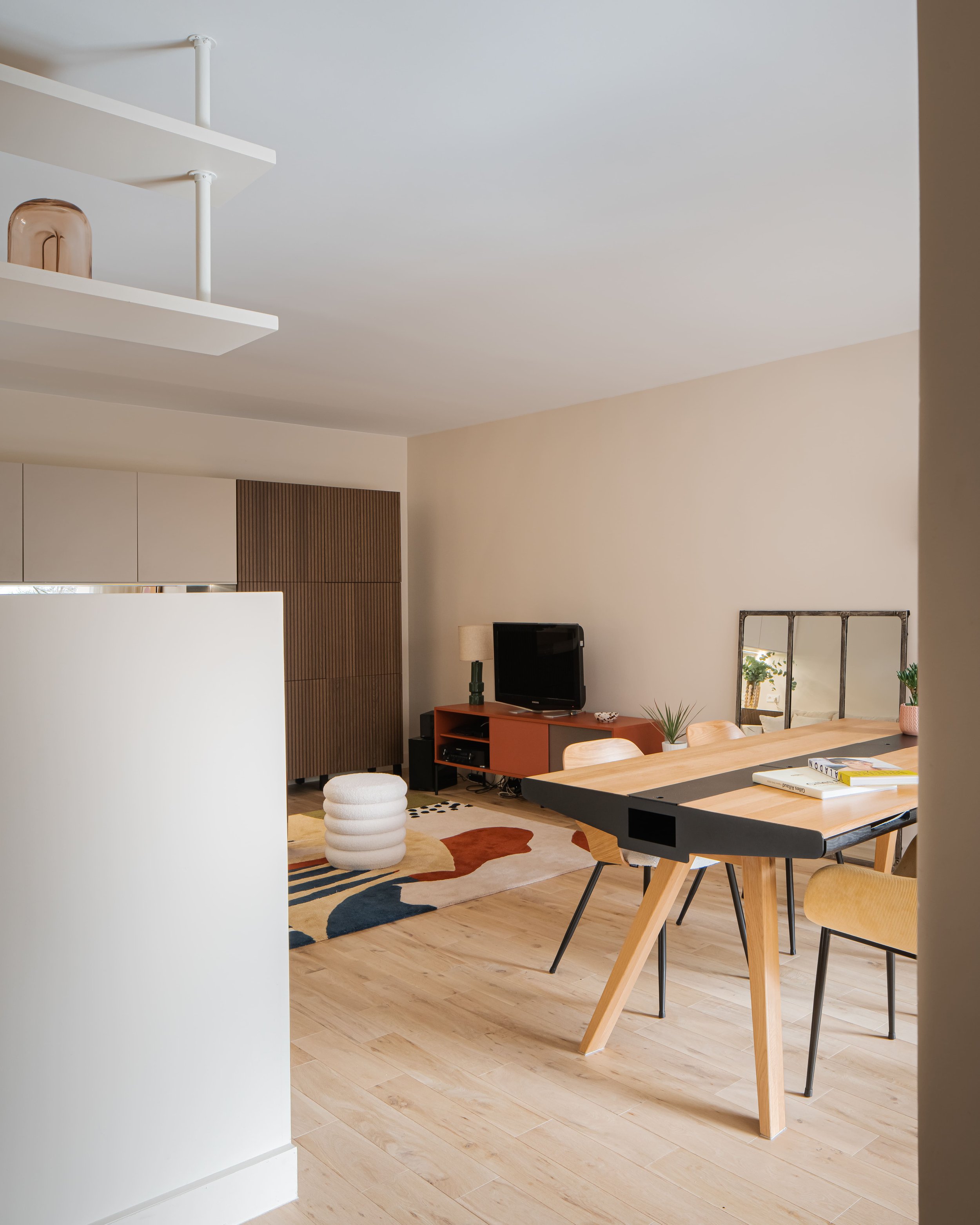 Intérieur d'un salon moderne avec un mur beige, un tapis coloré, une table en bois avec des chaises et une petite télévision sur un meuble orange.