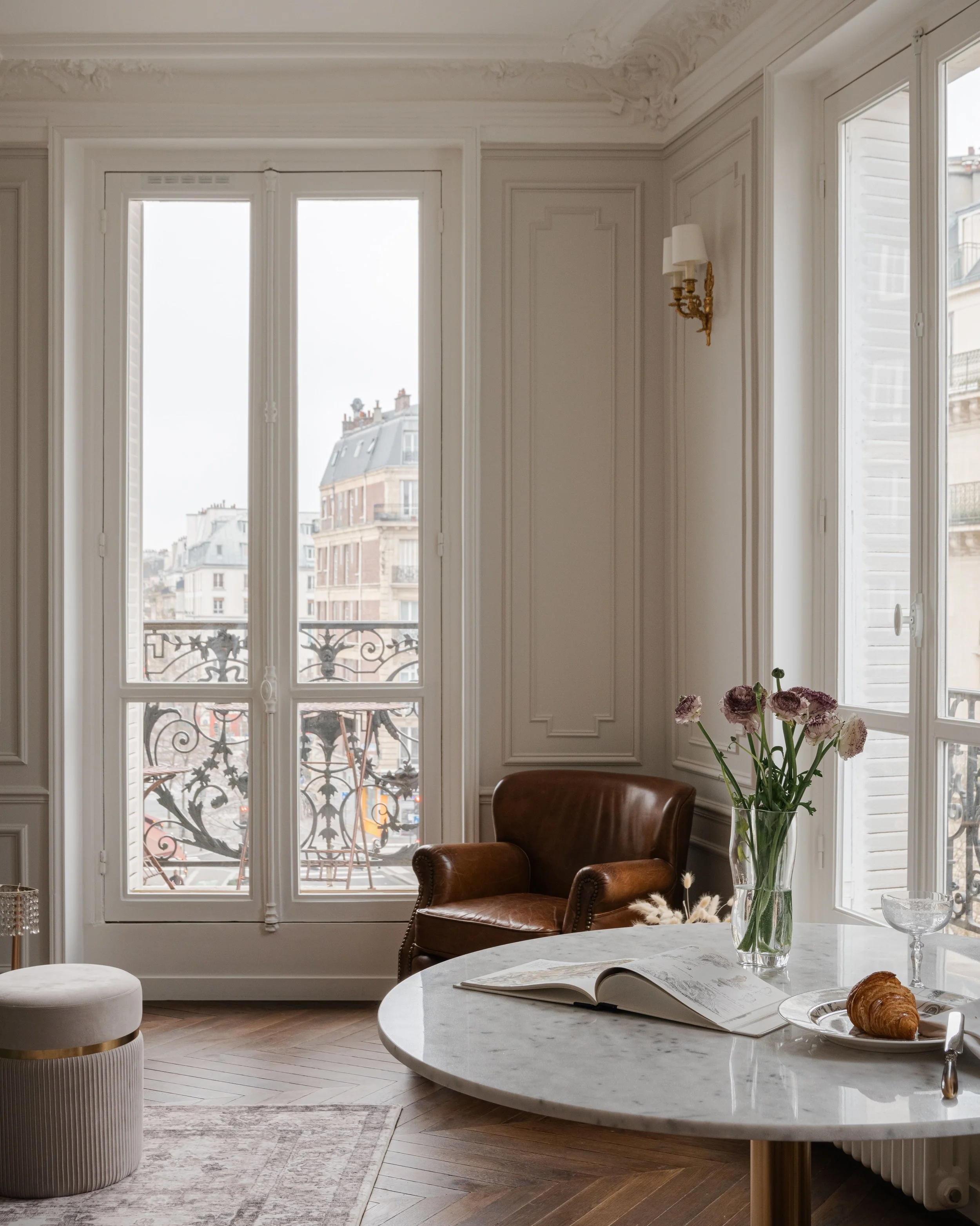 Salon lumineux avec fenêtres Parisiennes, table en marbre, vase de fleurs, livre ouvert, croissants, fauteuil en cuir brun, décoration élégante.
