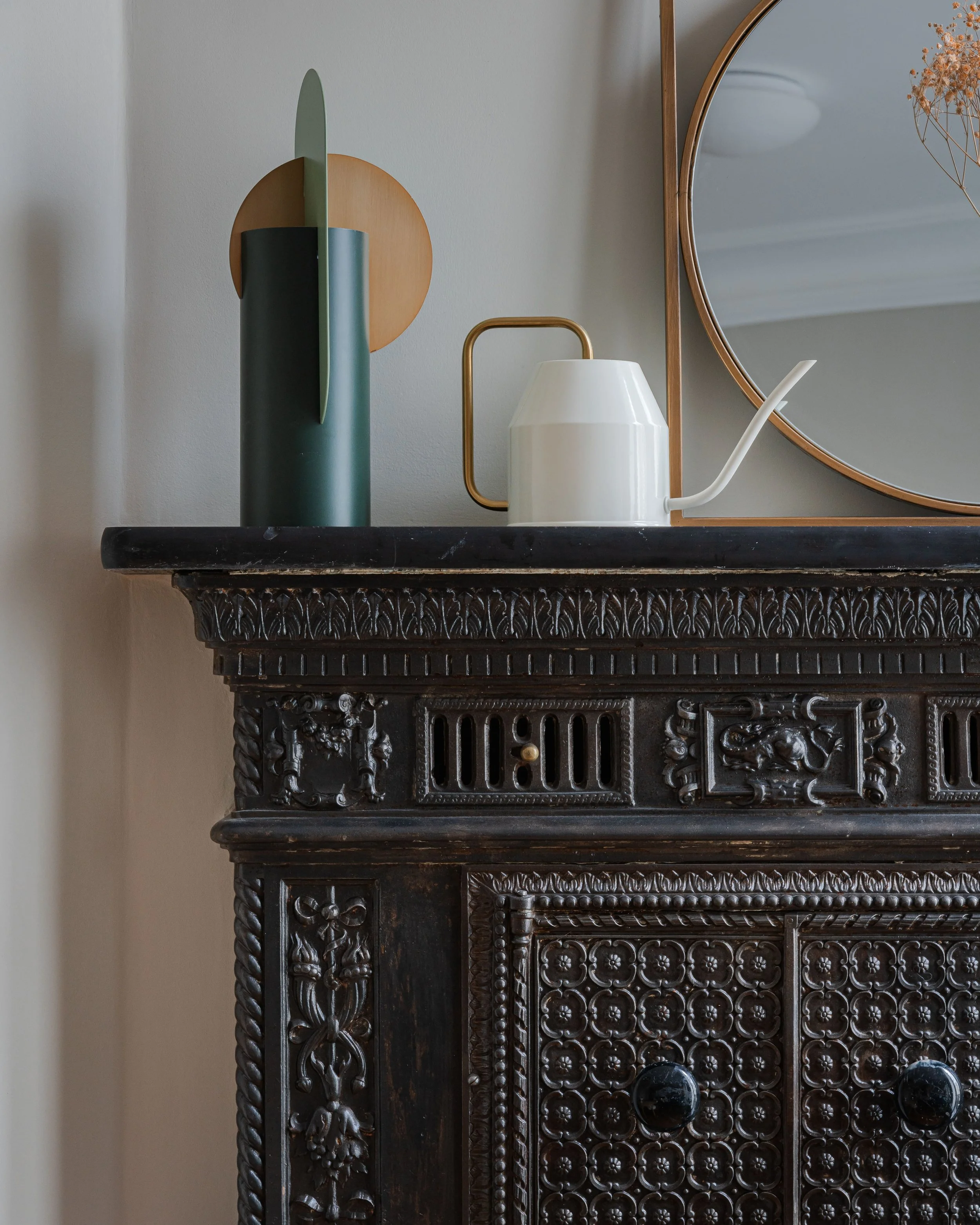 Une cheminée ancienne en bois sombre, décorée de motifs sculptés, avec un miroir rond en cadre doré au-dessus. Sur le dessus, une vase verte abstraite avec un cercle en bois derrière, un pot de style café avec un manche en or et un long bec verseur b