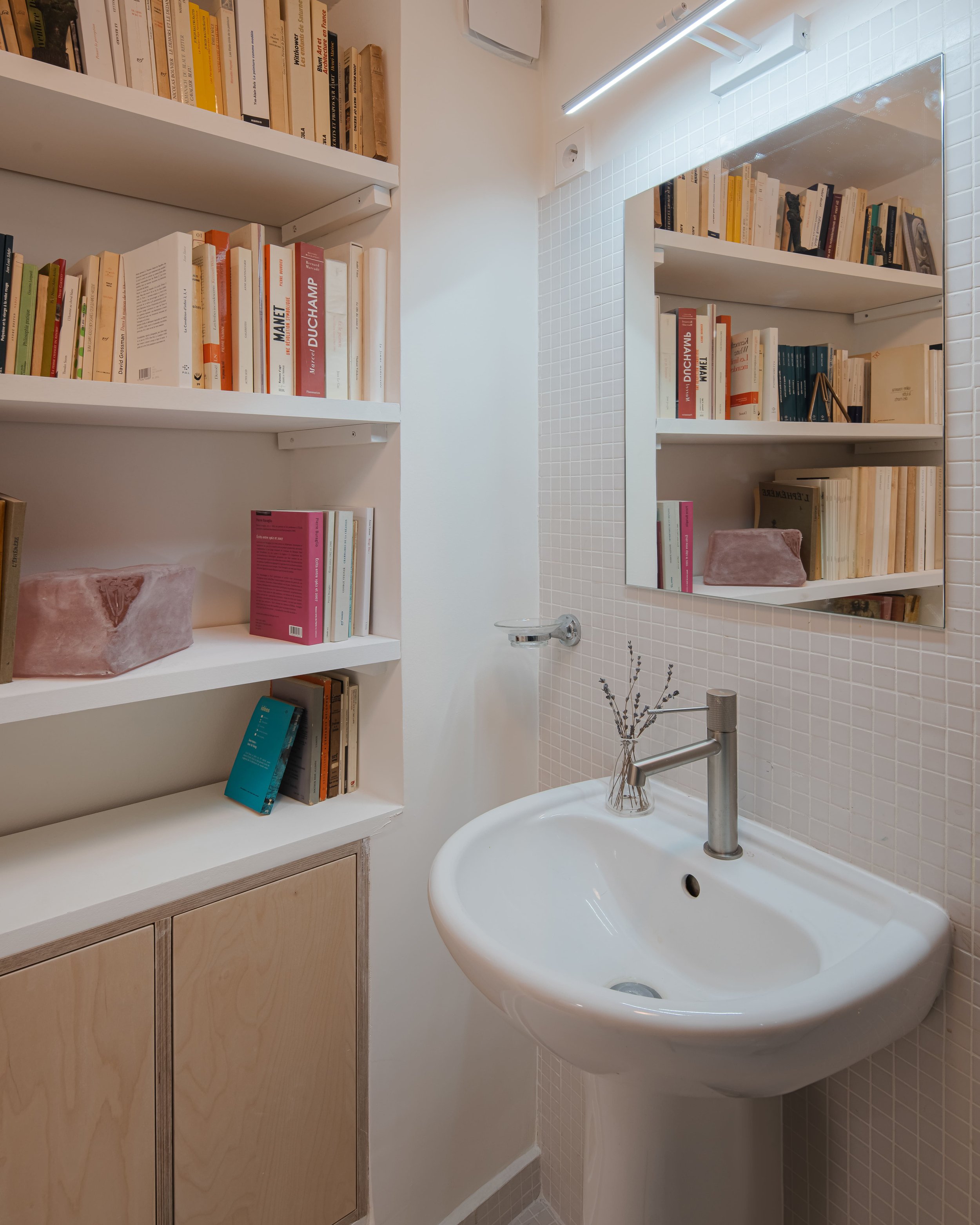Petite salle de bain avec un miroir rectangulaire, un évier blanc, un porte-savon en verre, un robinet en métal, et une petite étagère avec des livres et une brosse en pierre rose