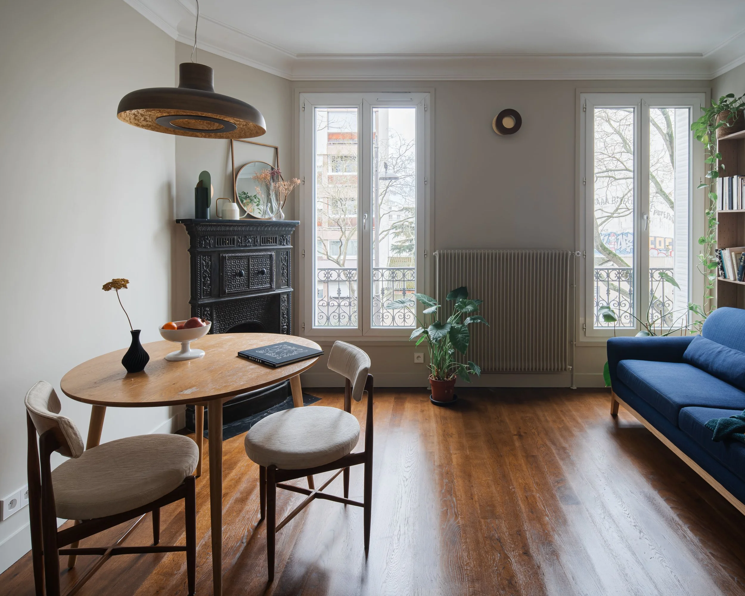 Intérieur d'un salon lumineux avec un tapis de bois, un canapé bleu, une table en bois avec trois chaises, une cheminée noire, des plantes vertes, et de grandes fenêtres laissant entrer la lumière naturelle.