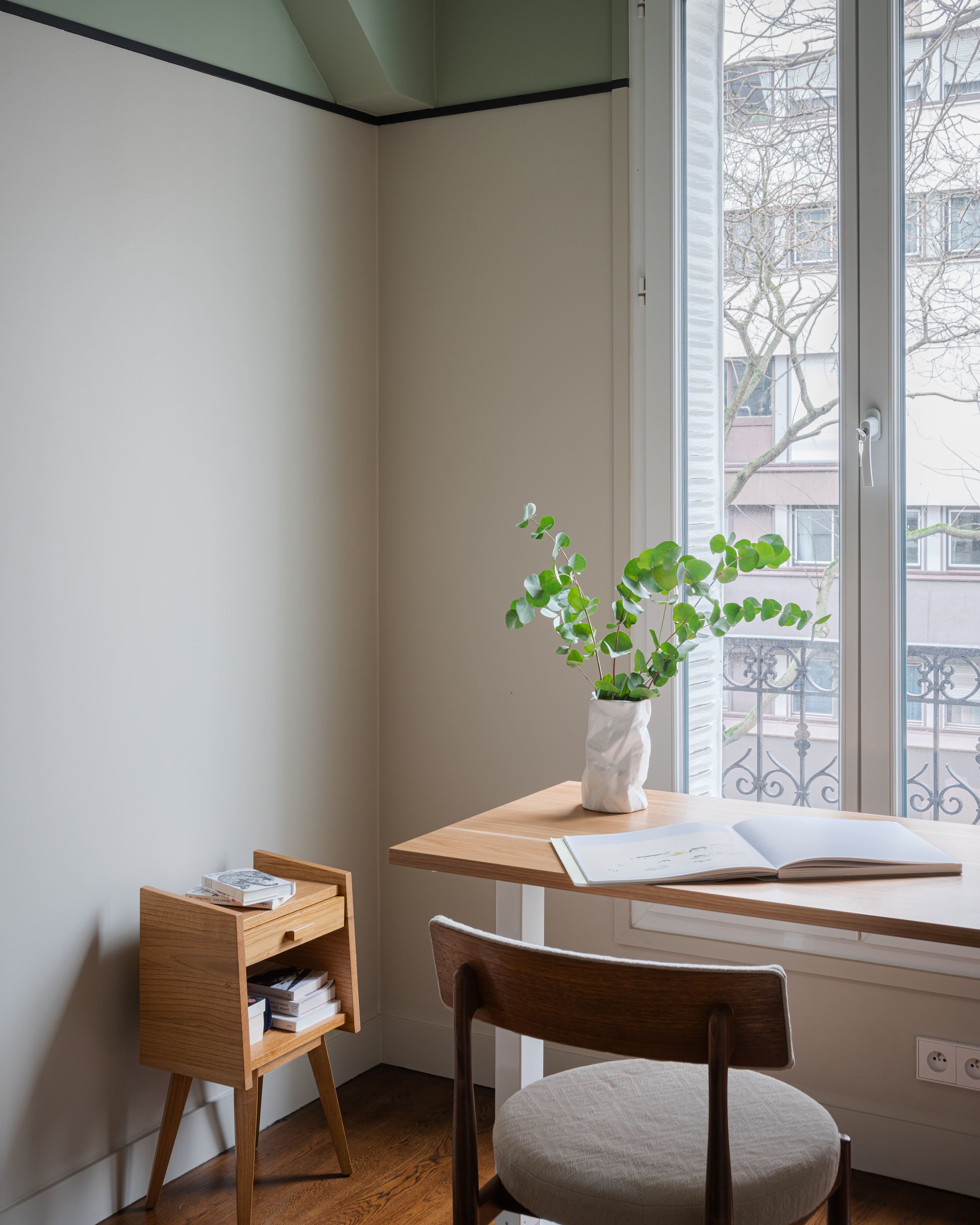 Intérieur d'un bureau avec une table en bois, une chaise en bois avec coussin clair, une plante verte dans un vase blanc, un livre ouvert, près d'une fenêtre donnant sur un bâtiment et des arbres nus, avec meubles en bois clair.