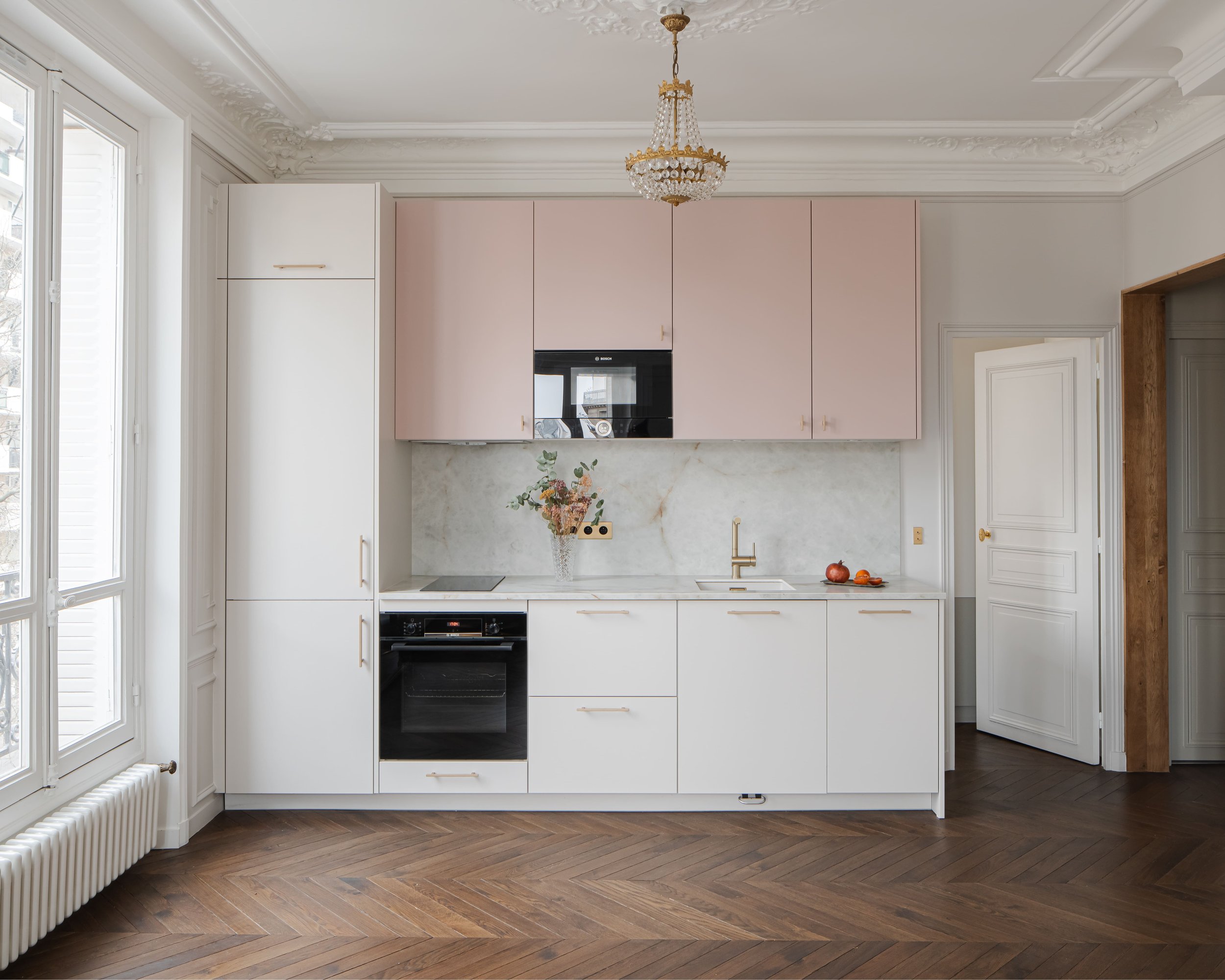 Cuisine élégante avec armoires blanches et rose pâle, plan de travail en marbre, chandelier suspendu, grande fenêtre avec radiateur en dessous, et sol en parquet herringbone.