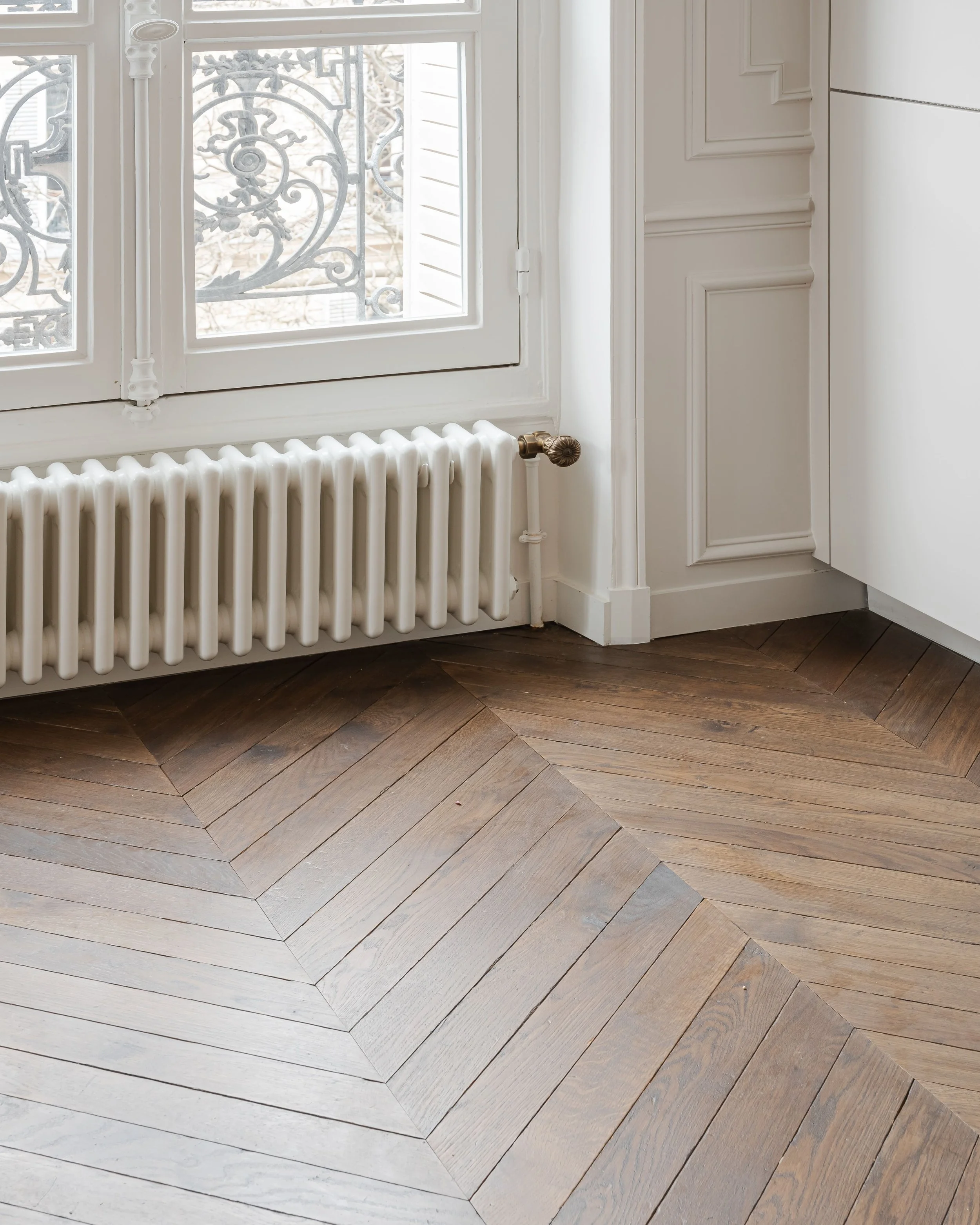 Fenêtre avec grille en métal, radiateur blanc en dessous, plancher en parquet en chevrons, et meubles blancs dans une pièce lumineuse.