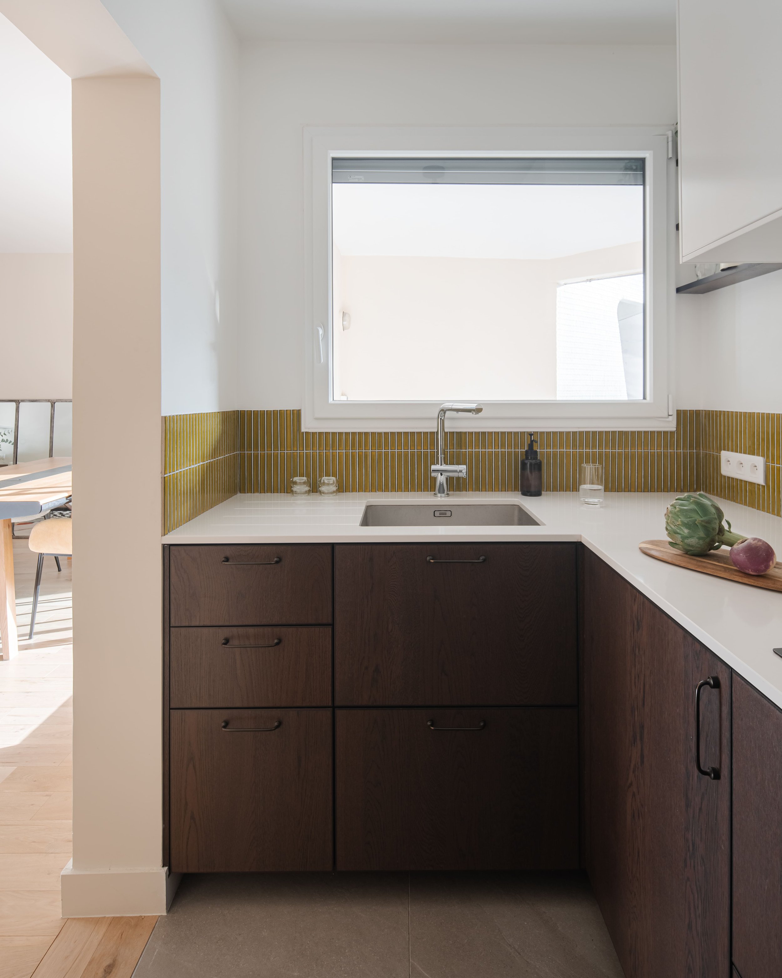 Coin cuisine avec évier, fenêtre au-dessus, comptoir avec un artichaut et légumes, cabines en bois foncé, dosseret en carrelage jaune, prise électrique sur le mur.