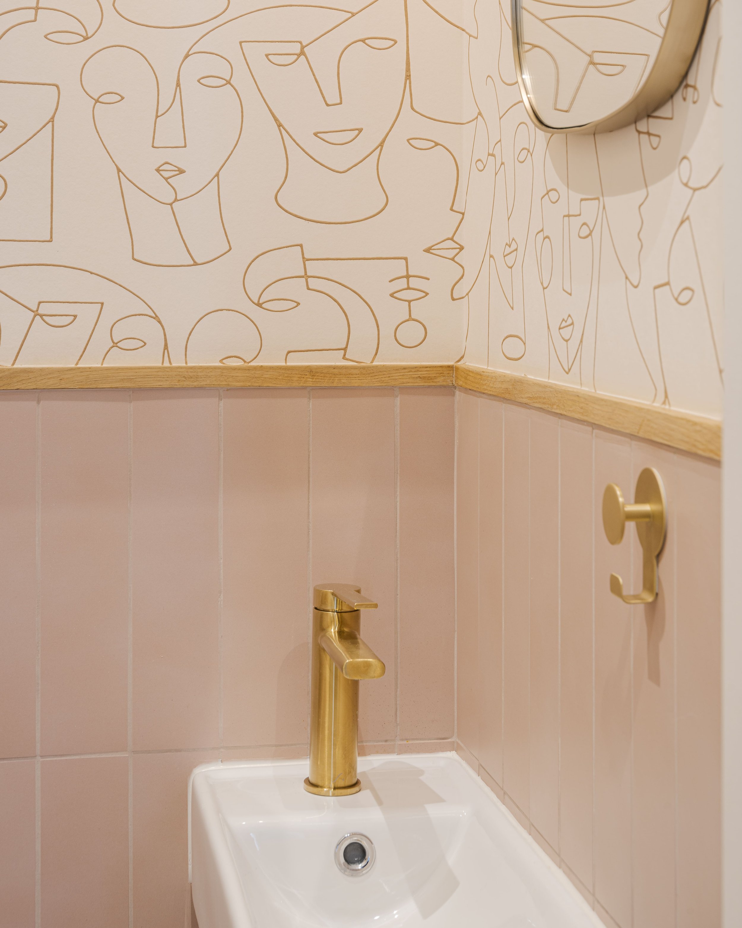 Un lavabo avec un robinet en or, un miroir et un mur décoré de dessins de visages féminins en ligne continue, tons beige et rose pastel.