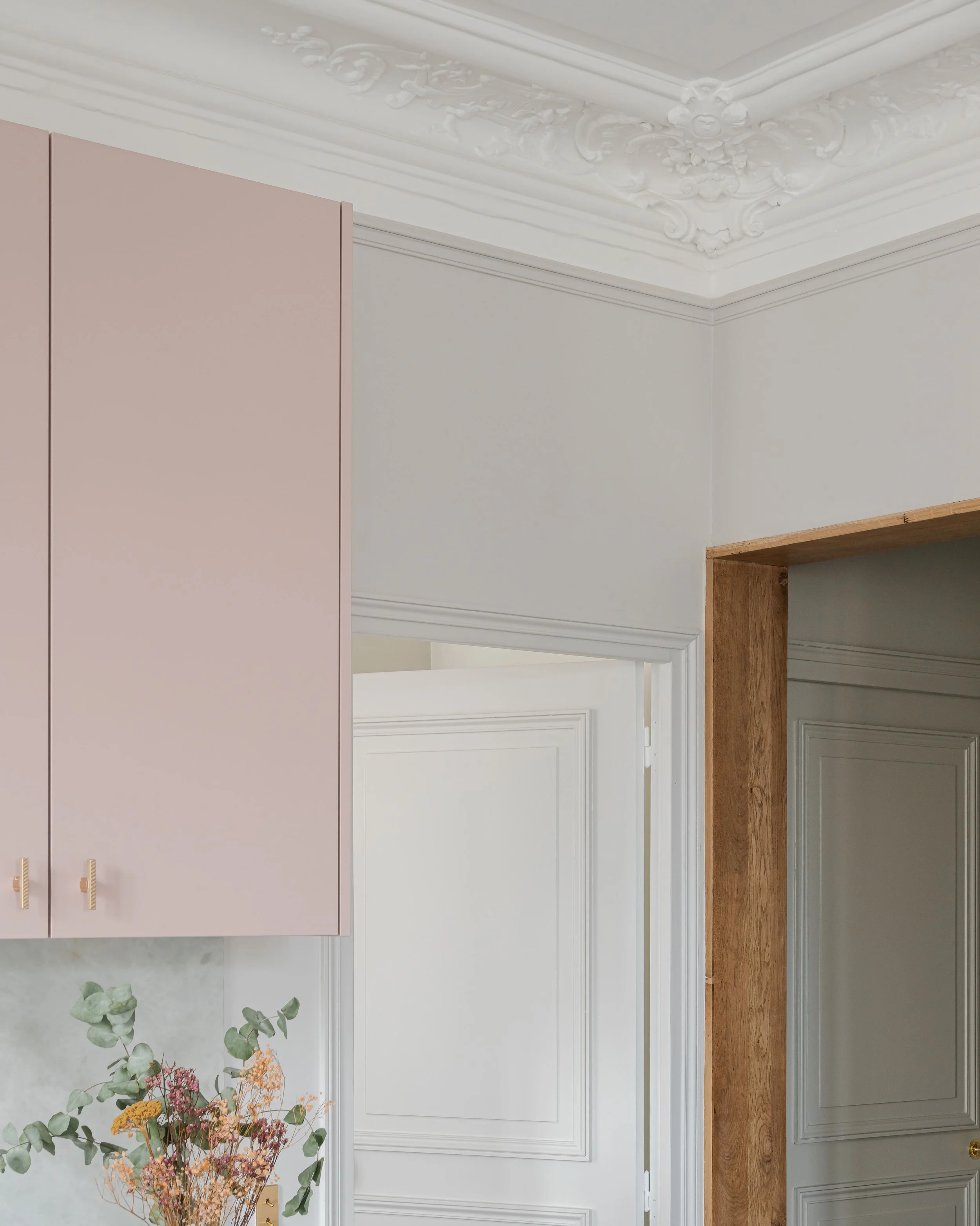 Vue partielle d'une cuisine élégante avec un placard rose pâle, un mur blanc, et une arche en bois massif.