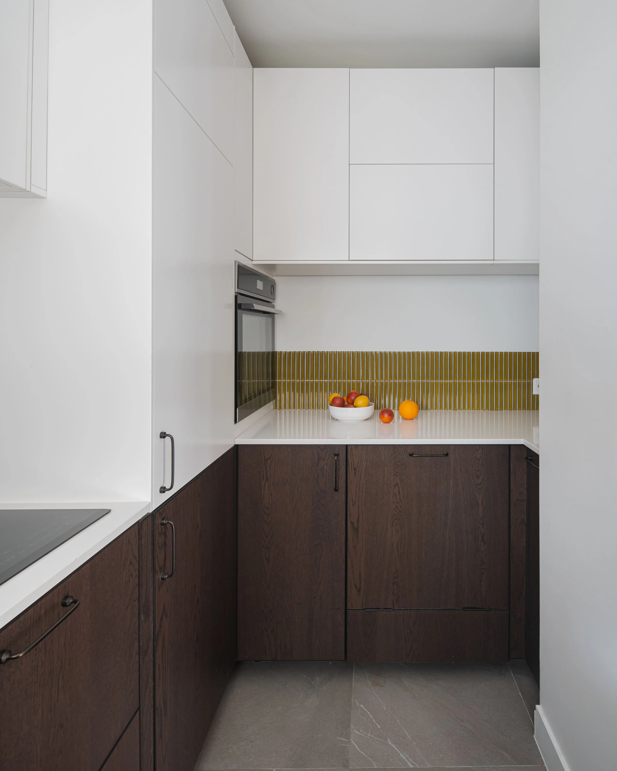 Coin de cuisine moderne avec armoires blanches et bois foncé, comptoir blanc, des fruits dans un bol, et un dosseret en petits carreaux jaunes