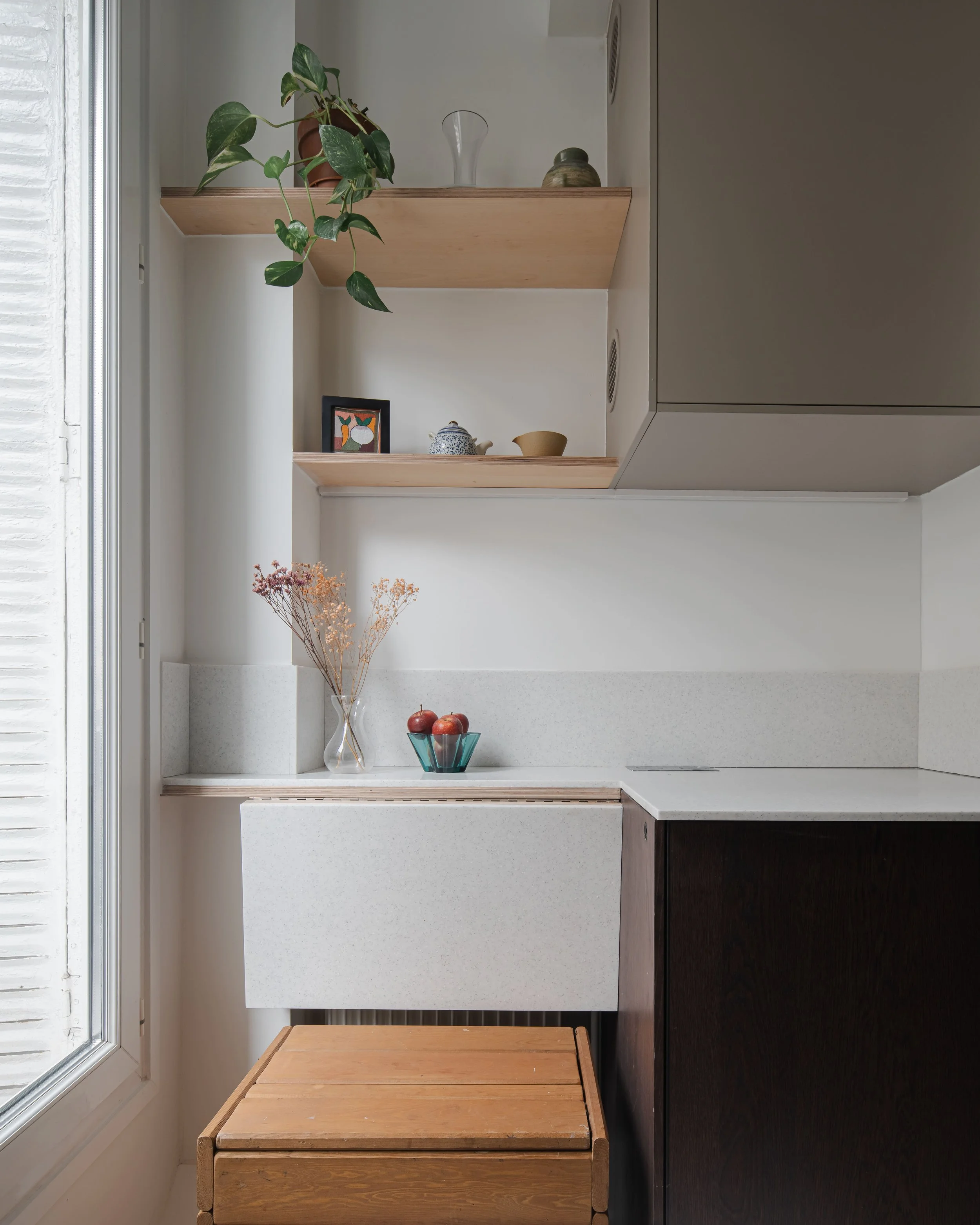 Cuisine minimaliste avec étagères en bois, plantes vertes, vaisselle, un vase avec des fleurs séchées, un bol de fruits, et un meuble en bois.