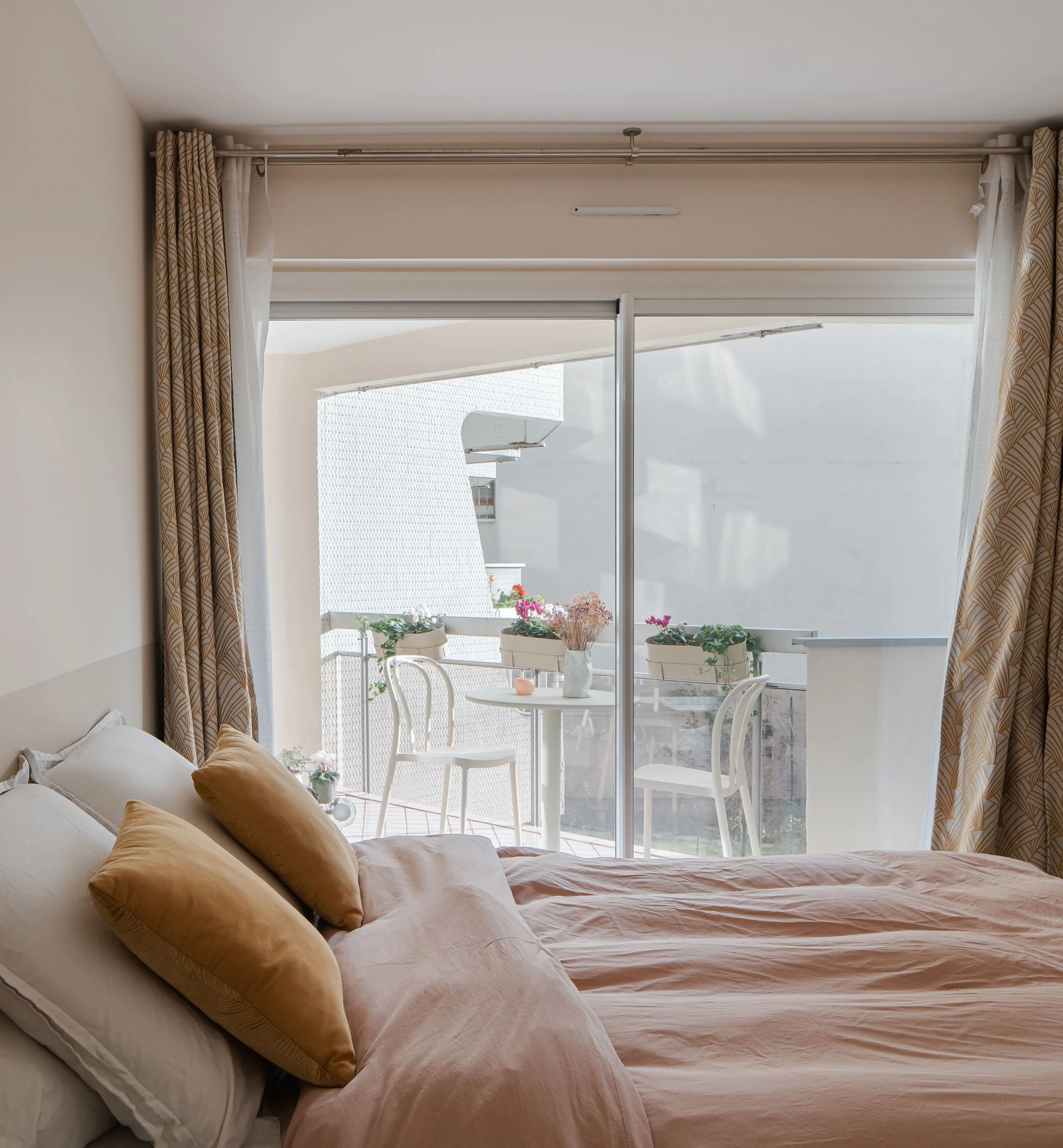 Chambre avec lit rose et vue sur un balcon avec table, chaises et plantes. Transparents avec rideaux beige avec motifs.