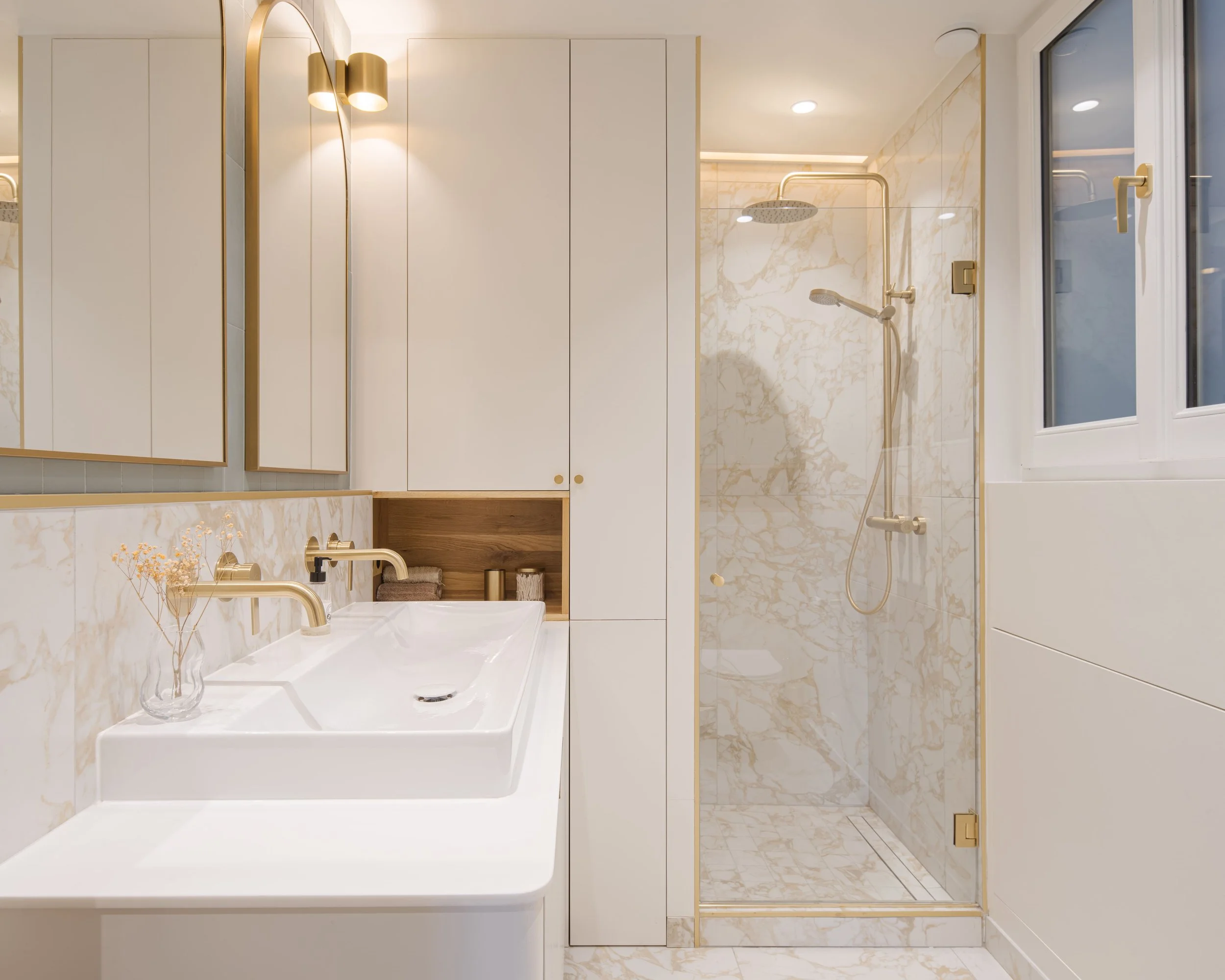 Salle de bain moderne avec douche en marbre, lavabo blanc, meubles blancs, miroir avec éclairage doré, et fenêtre