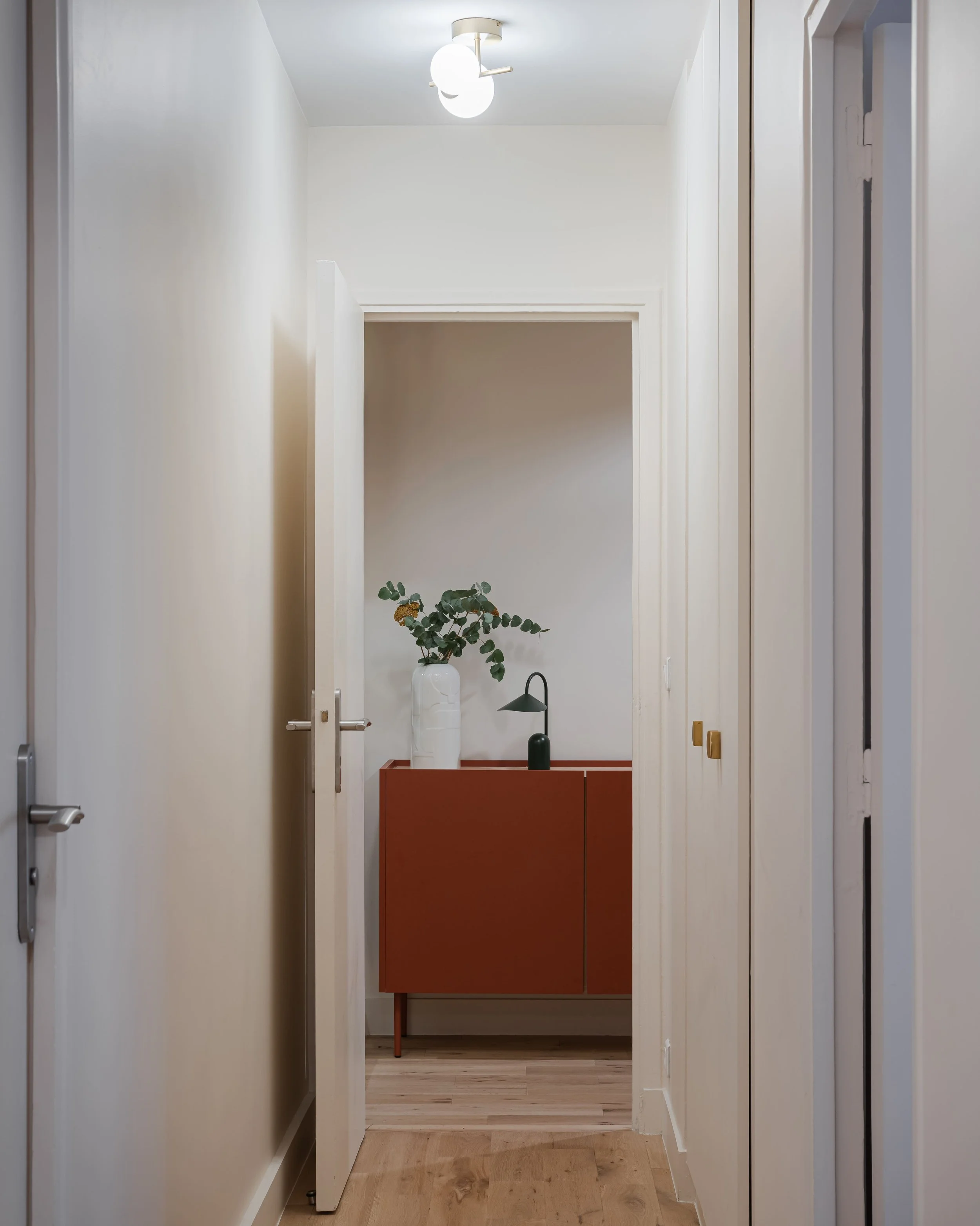 Couloir avec murs clairs et porte entrouverte menant à un meuble rouge avec une plante et une lampe noire dessus.