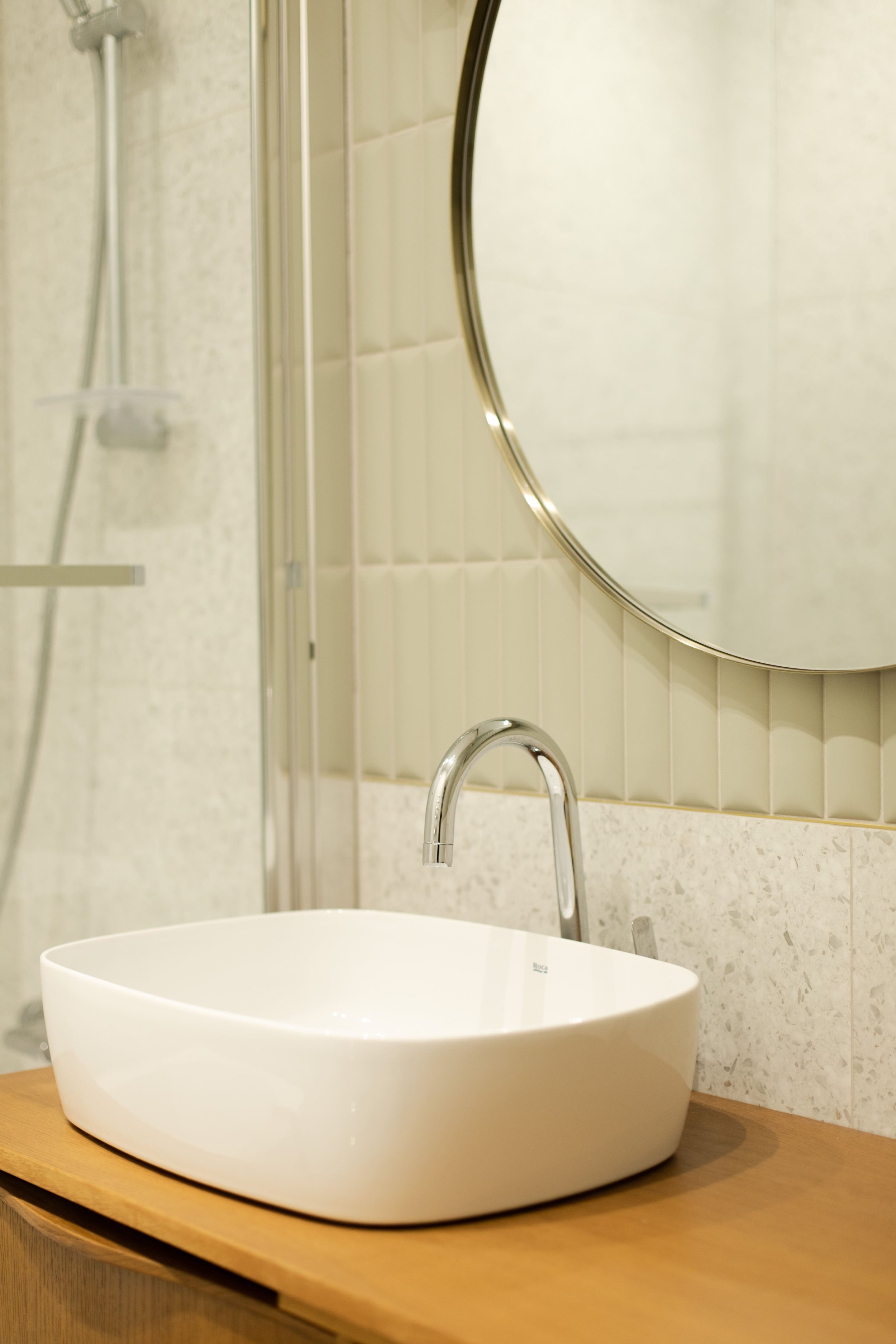 Lavabo blanc ovale en céramique sur une étagère en bois dans une salle de bain. Miroir rond au-dessus. Douche visible à gauche.