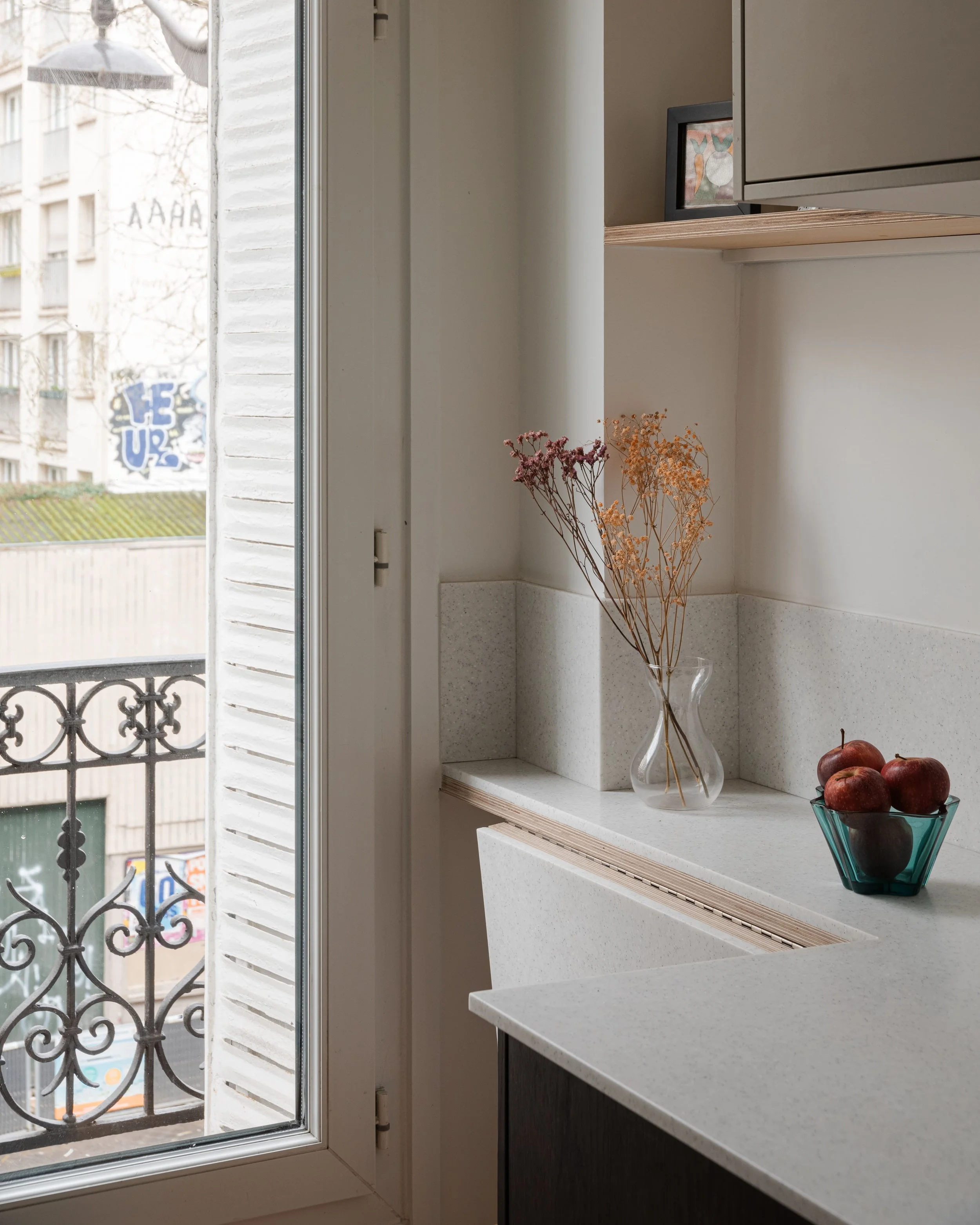 Intérieur d'une cuisine avec une fenêtre ouverte donnant sur un balcon en fer forgé, décorée avec un vase contenant des fleurs sèches et un bol de pommes rouges.