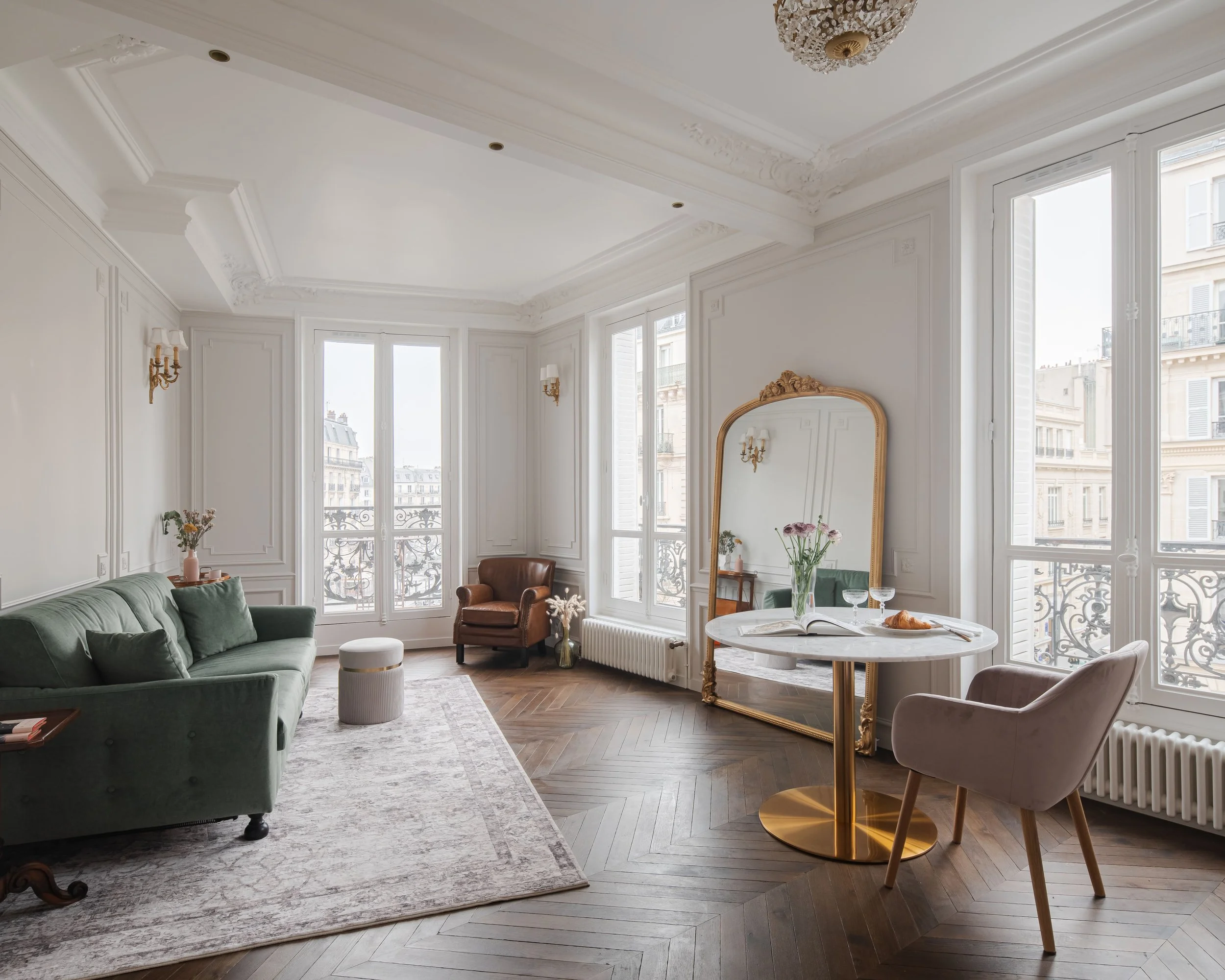Salon intérieur élégant avec grand miroir, canapé vert, fauteuil en cuir marron, table ronde avec bouquet de fleurs, grande fenêtres avec vue sur un immeuble parisien, parquet en bois.