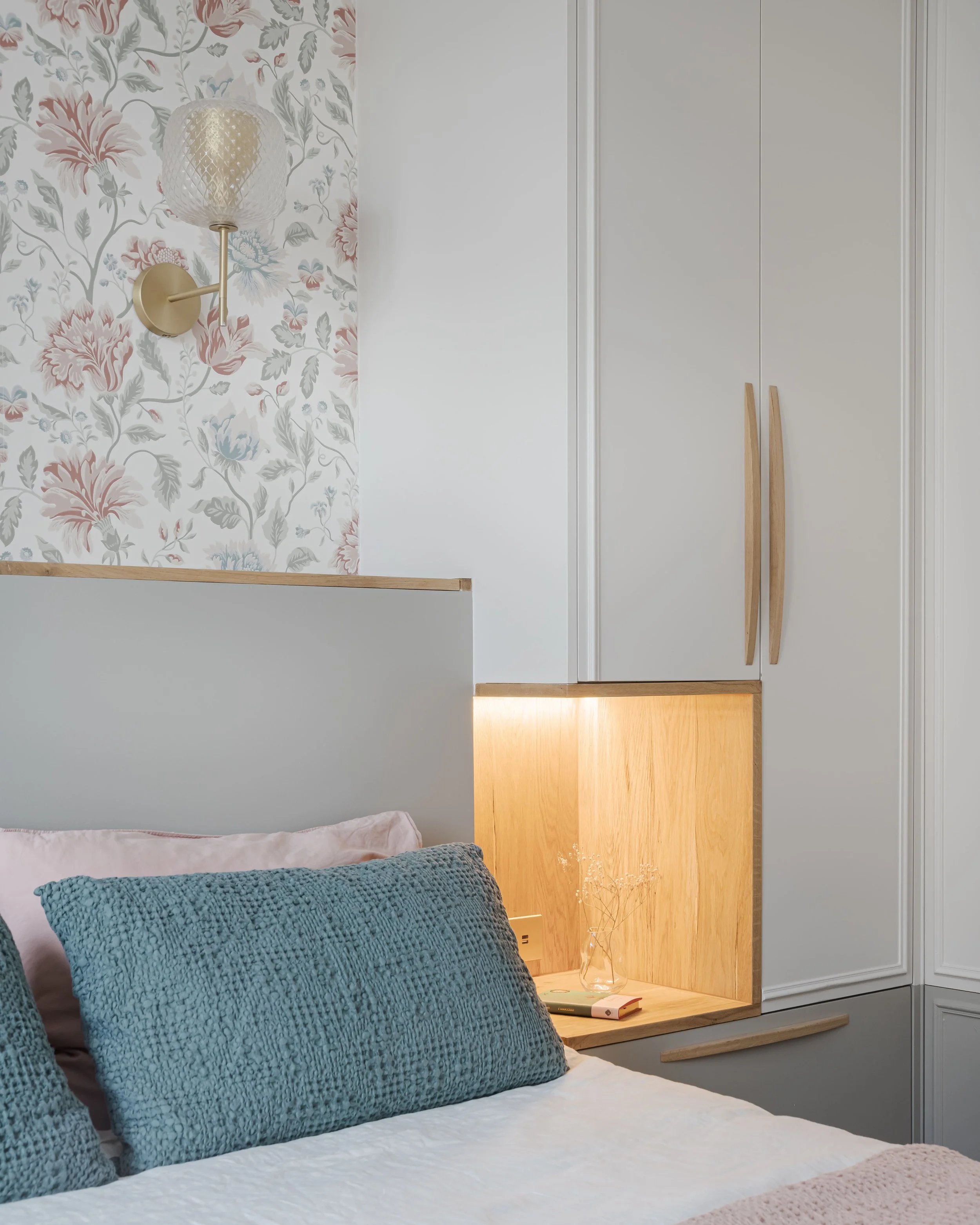 Coin de chambre avec lit, coussins bleus, lampadaire mural et armoire blanche avec poignées en bois, mur avec papier peint floral pastel.