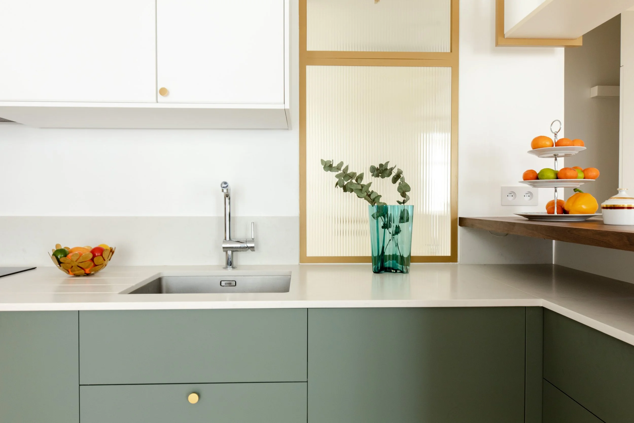 Cuisine moderne avec un évier, un bol de fruits, une plante verte dans un vase en verre bleu, et une table à thé ou dessert avec des fruits