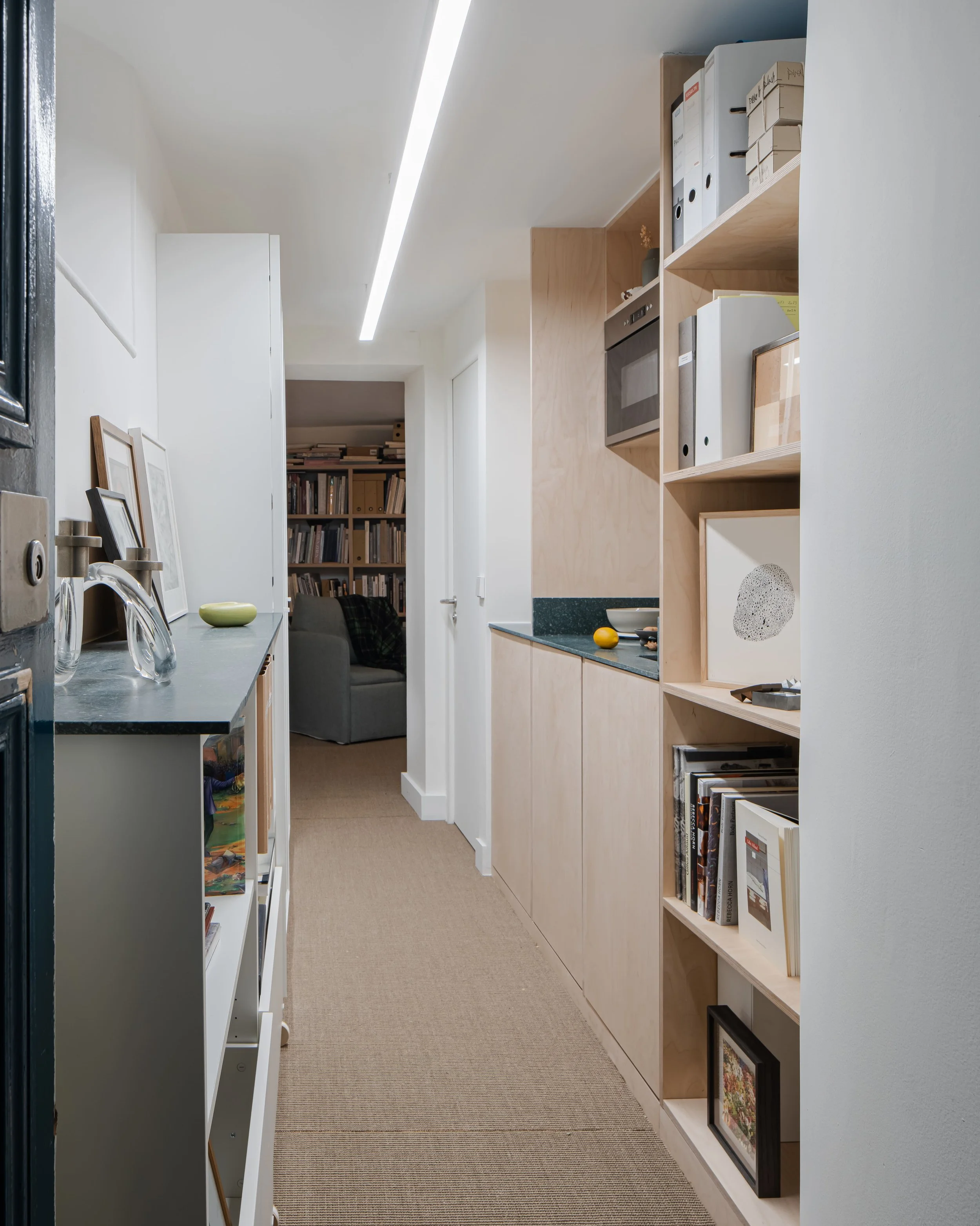 Un couloir étroit dans un appartement moderne avec étagères en bois contenant des livres, un four encastré, une table avec quelques objets décoratifs, et un espace salon visible au fond avec une bibliothèque pleine de livres.