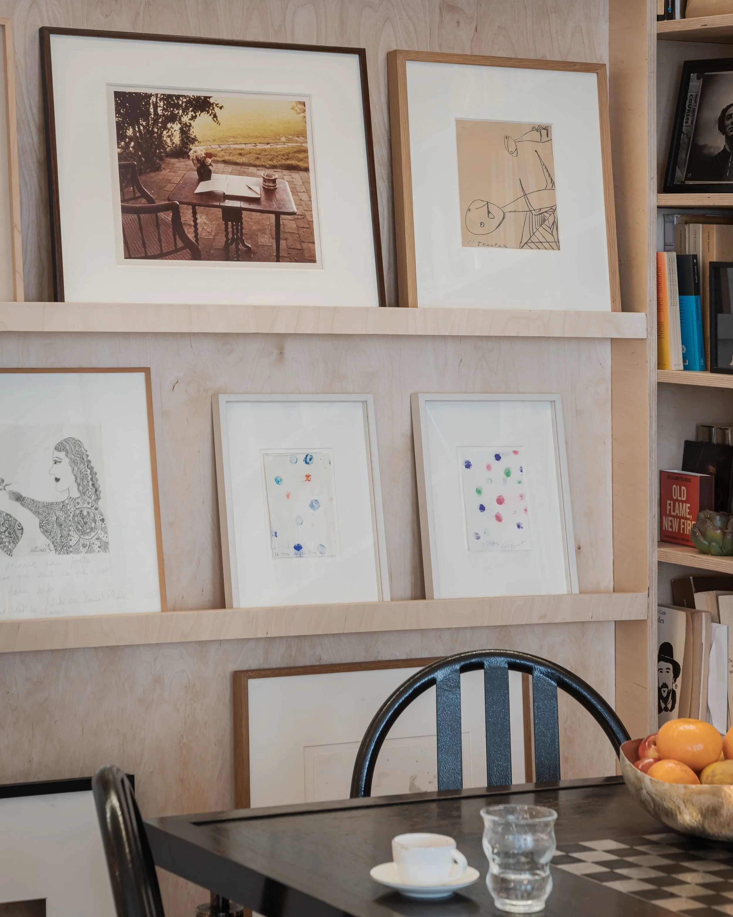 Une étagère avec plusieurs tableaux, dont un photo d'une table en plein air au coucher du soleil, des dessins et des œuvres d'art abstraites. À droite, des livres et une plante décorative, en partie visible. Au premier plan, une table avec une tasse,