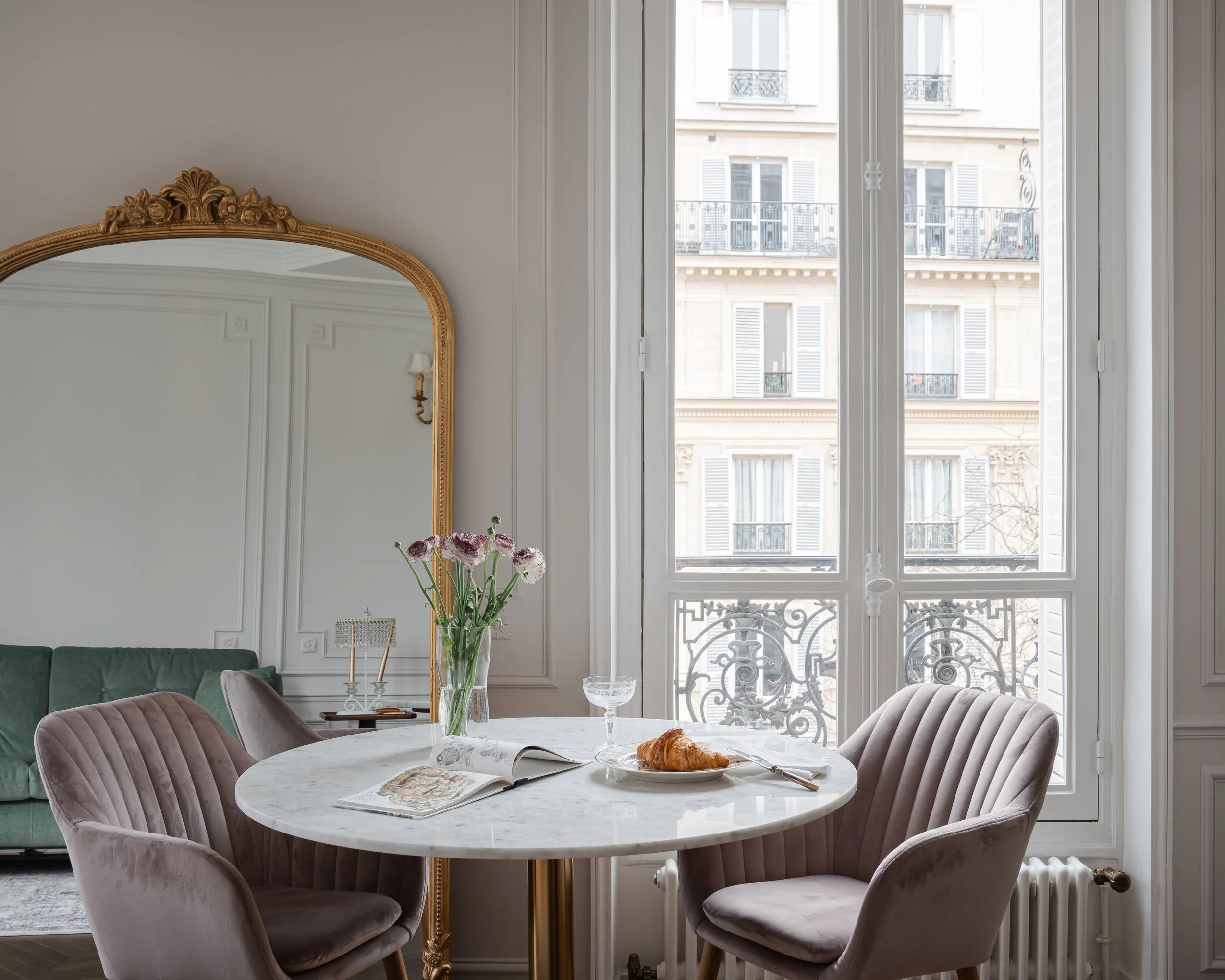 Salle à manger élégante avec table en marbre blanc, chaises en velours rose, une fleur dans un vase, un croissant sur une assiette, et un grand miroir doré, devant une fenêtre donnant sur un bâtiment parisien.