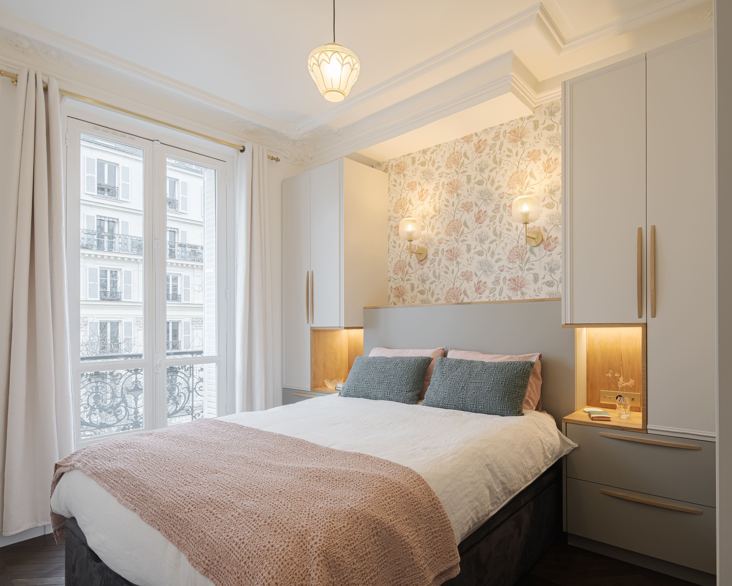 Chambre à coucher moderne avec lit double, draps blancs, coussins gris, mur de papier peint floral derrière la tête de lit, deux lampes murales, grande fenêtre avec rideaux blancs et vue sur des immeubles parisiens.