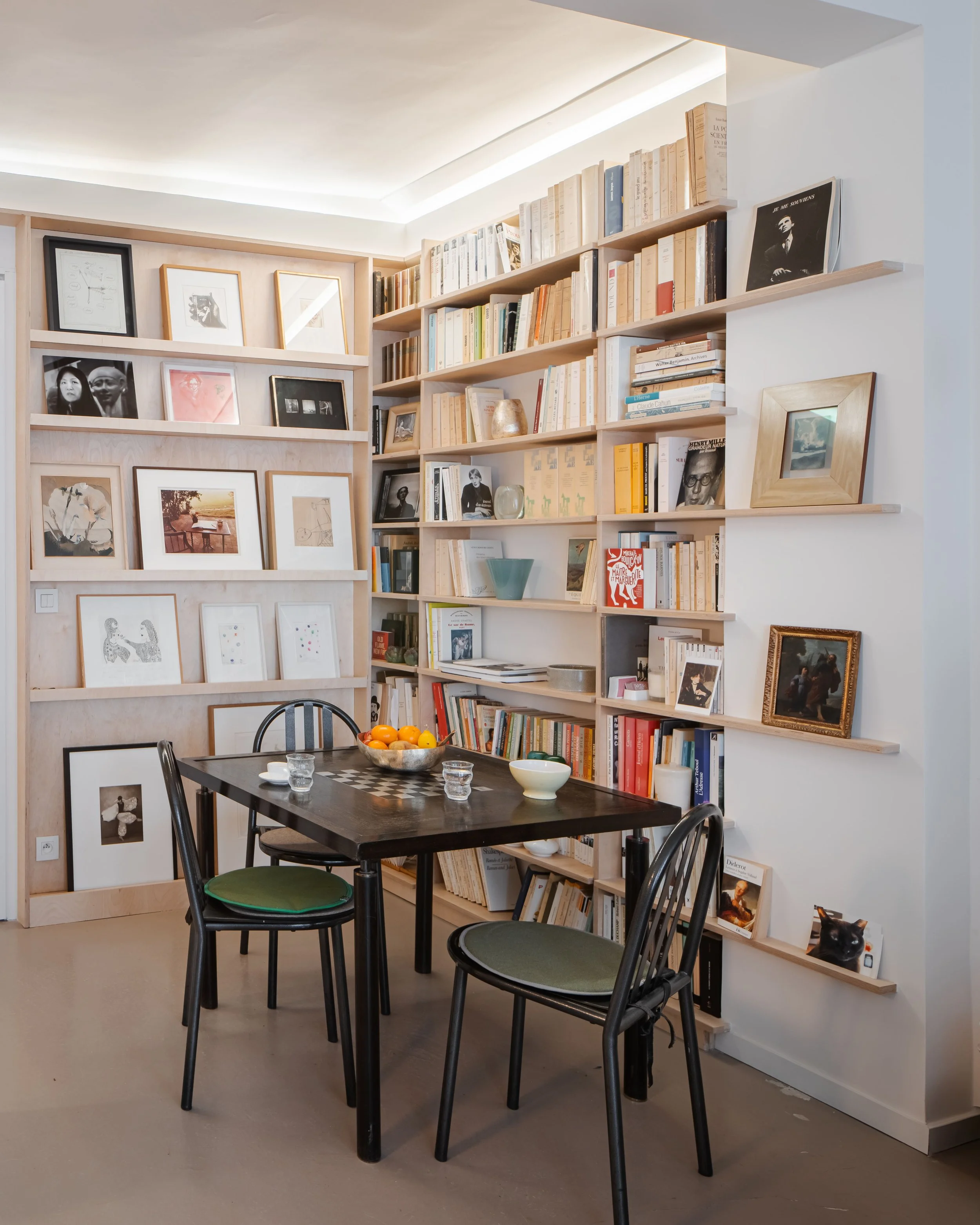 Salle à manger avec une table en bois noir, quatre chaises et étagères remplies de livres et de cadres sur le mur.