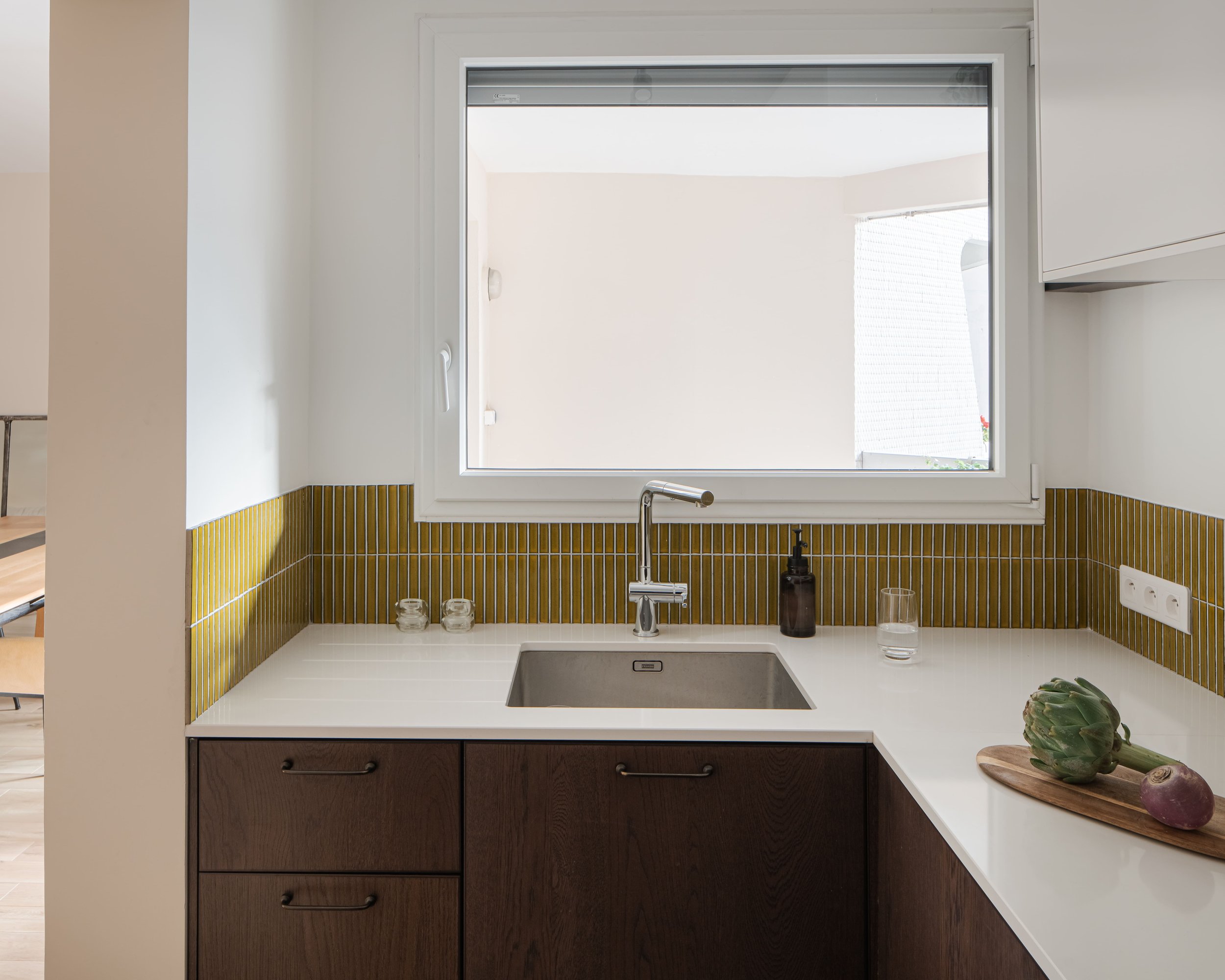 Cuisine avec fenêtre, évier en acier inoxydable, comptoir blanc, carrelage vert olive, dessus de comptoir avec artichaut, oignon, verre d'eau, savon et deux petits bocaux.