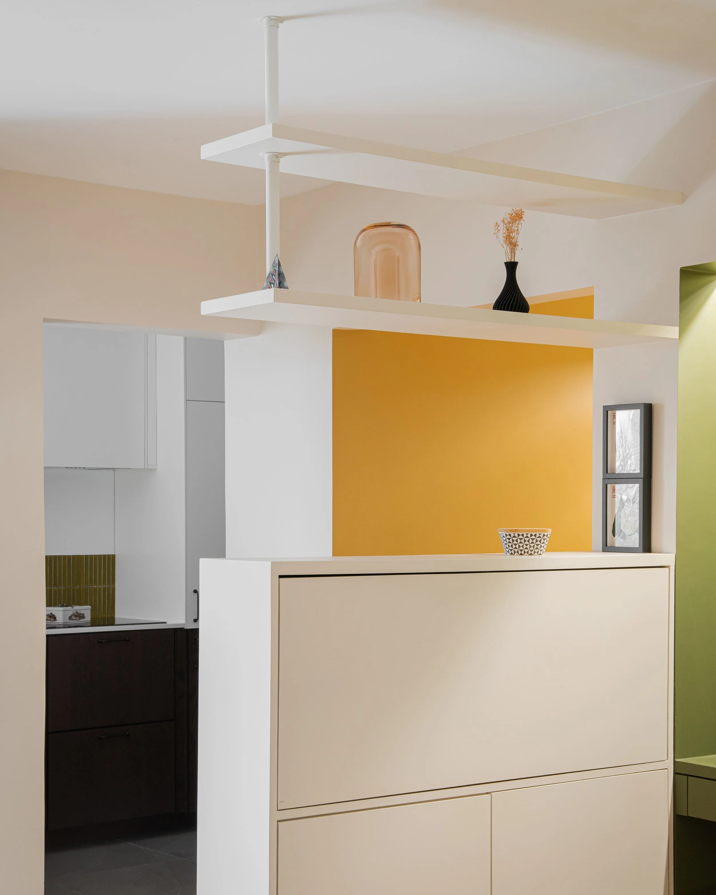 Décor intérieur moderne avec étagères blanches, murs peints en beige, jaune et vert, dans un espace ouvert avec éléments décoratifs comme vases et cadres.
