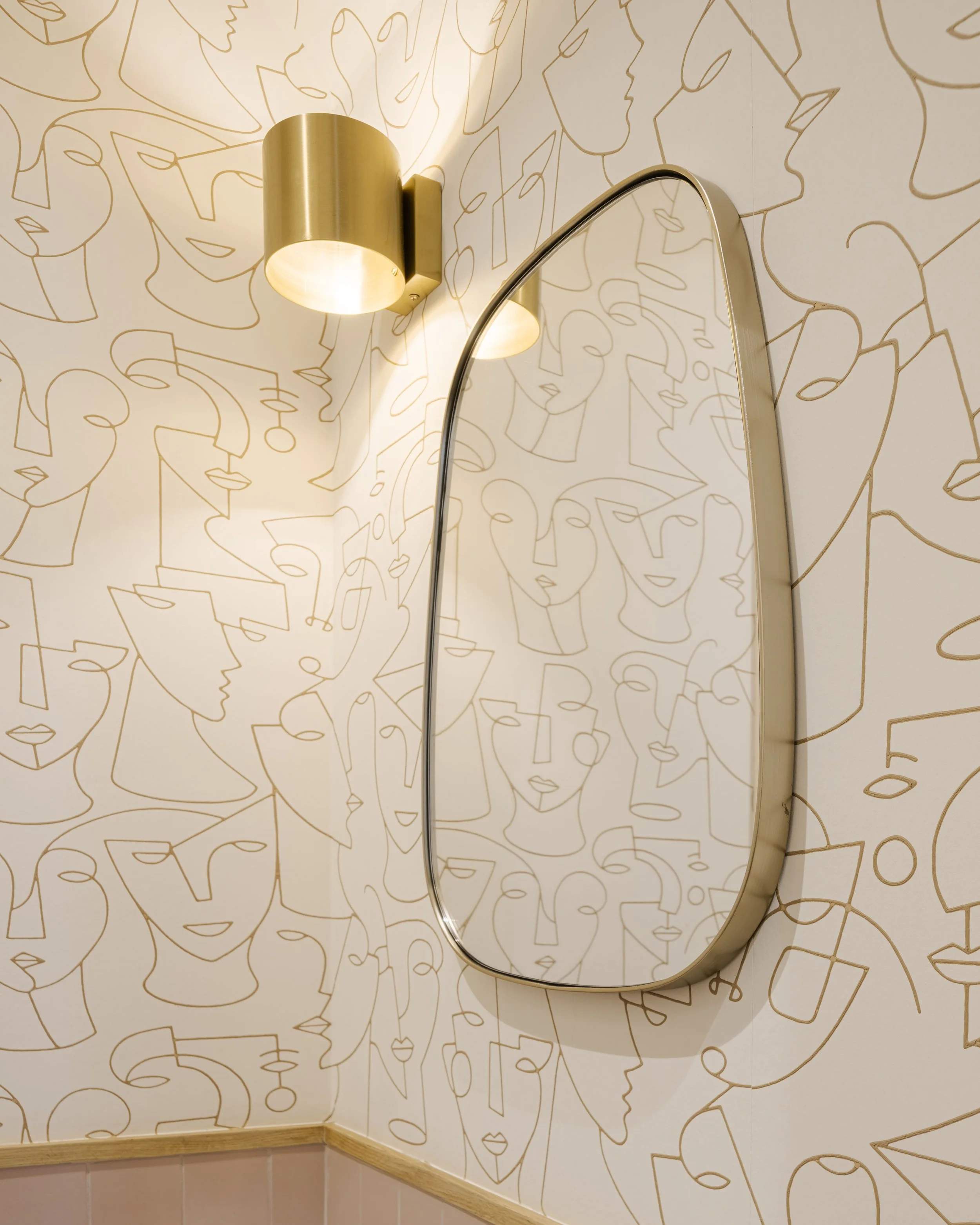Un miroir ovale avec un cadre doré, fixé à un mur décoré avec un motif de lignes dorées dessinant des visages abstraits, avec une applique murale dorée à gauche, éclairant la scène.