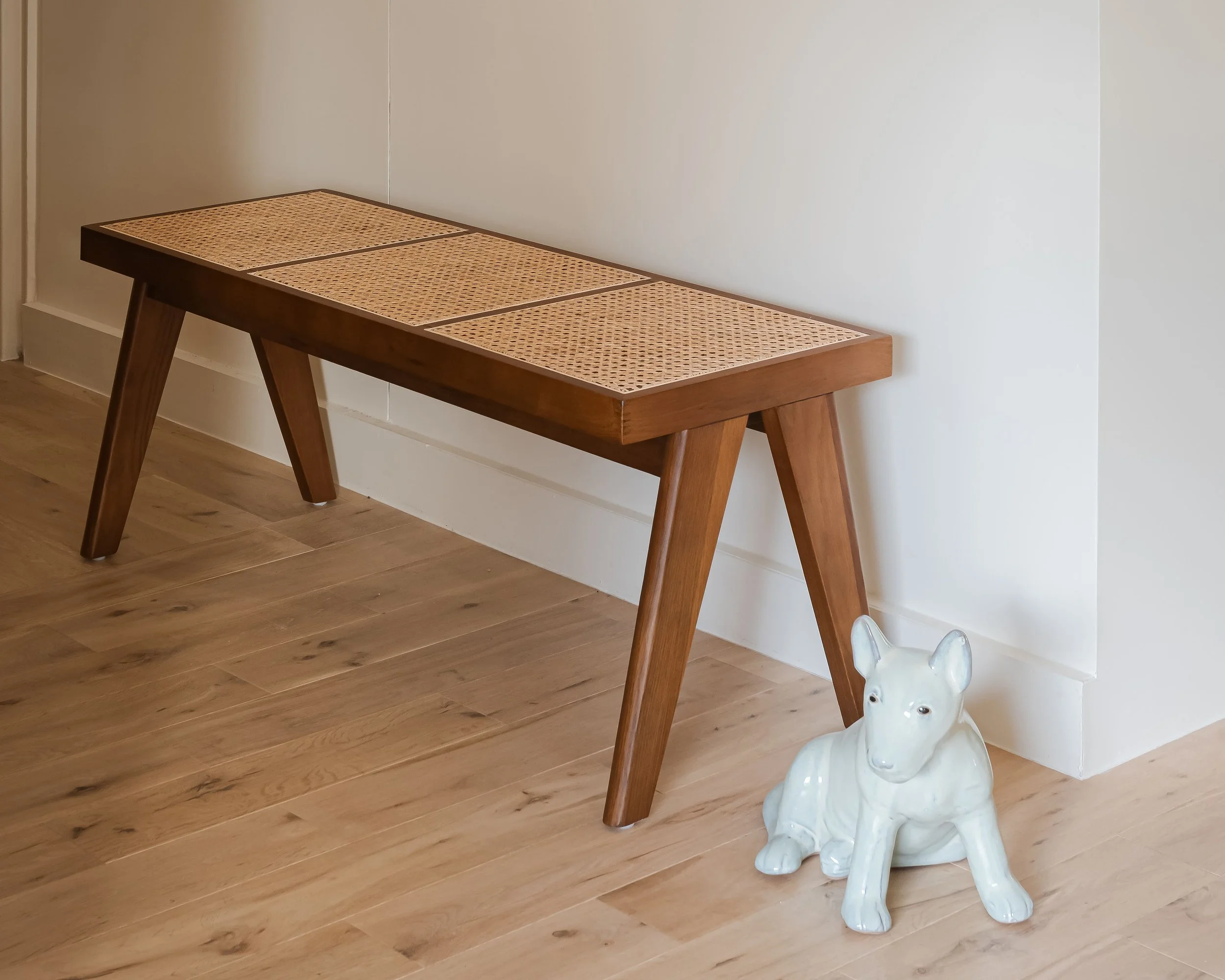 Un banc en bois avec assise en cannage, situé dans un intérieur avec plancher en bois clair, à côté d'une sculpture en céramique d'une petite famille de chiens